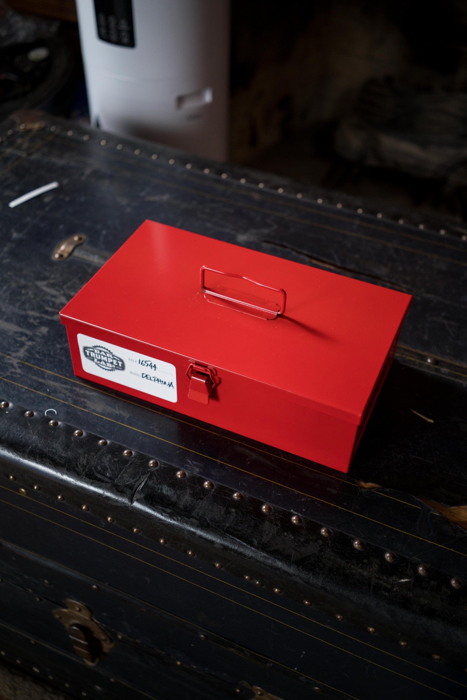 A bright red metal box with a handle and latch rests on a black, vintage trunk with brass studs. Beside it, an Ear Trumpet Labs Delphina Microphone stands out against the dimly lit background.