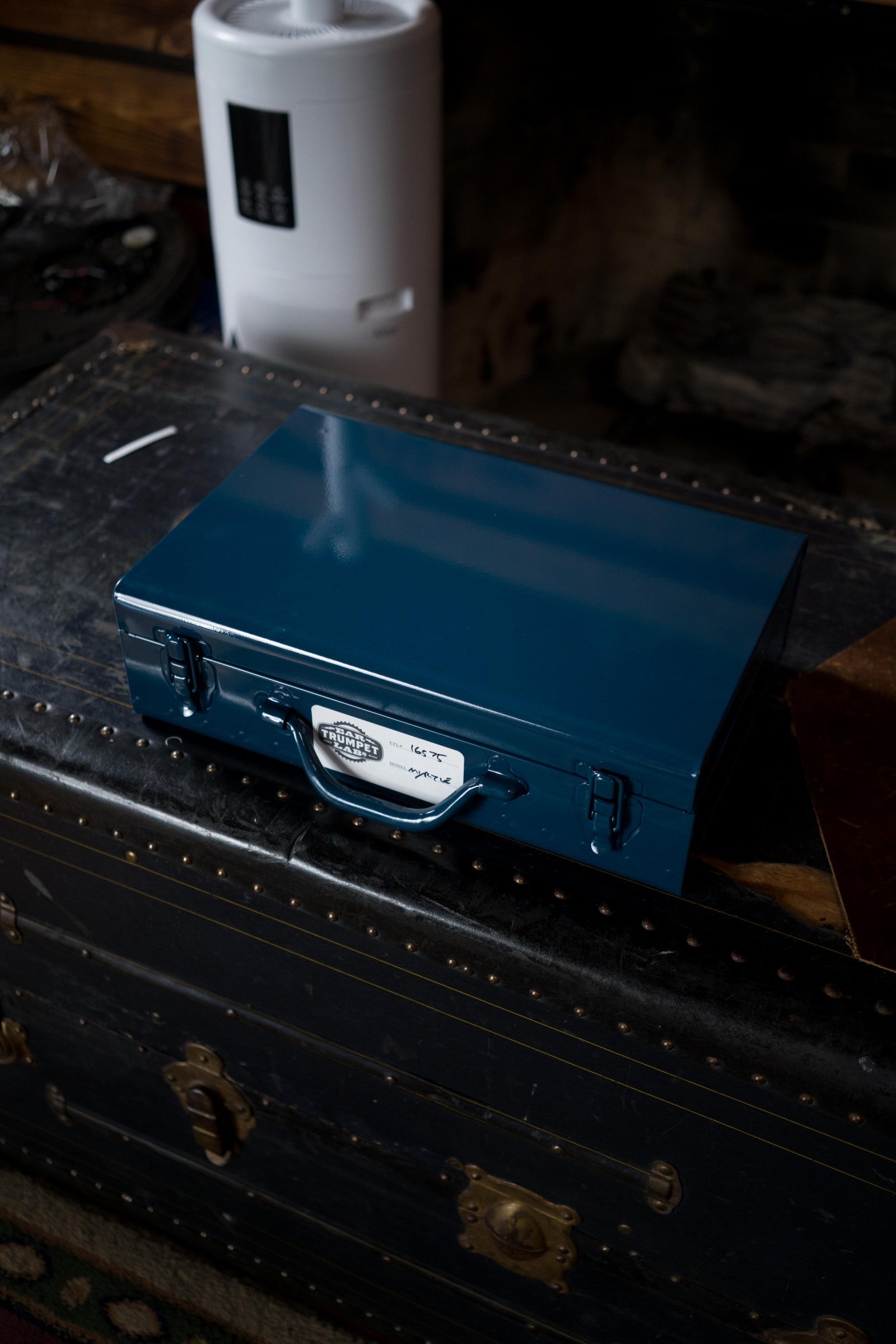 A closed blue metal cash box sits atop a black vintage trunk with metal studs in a dimly lit room. An Ear Trumpet Labs Myrtle Microphone and a white electronic device appear in the background.