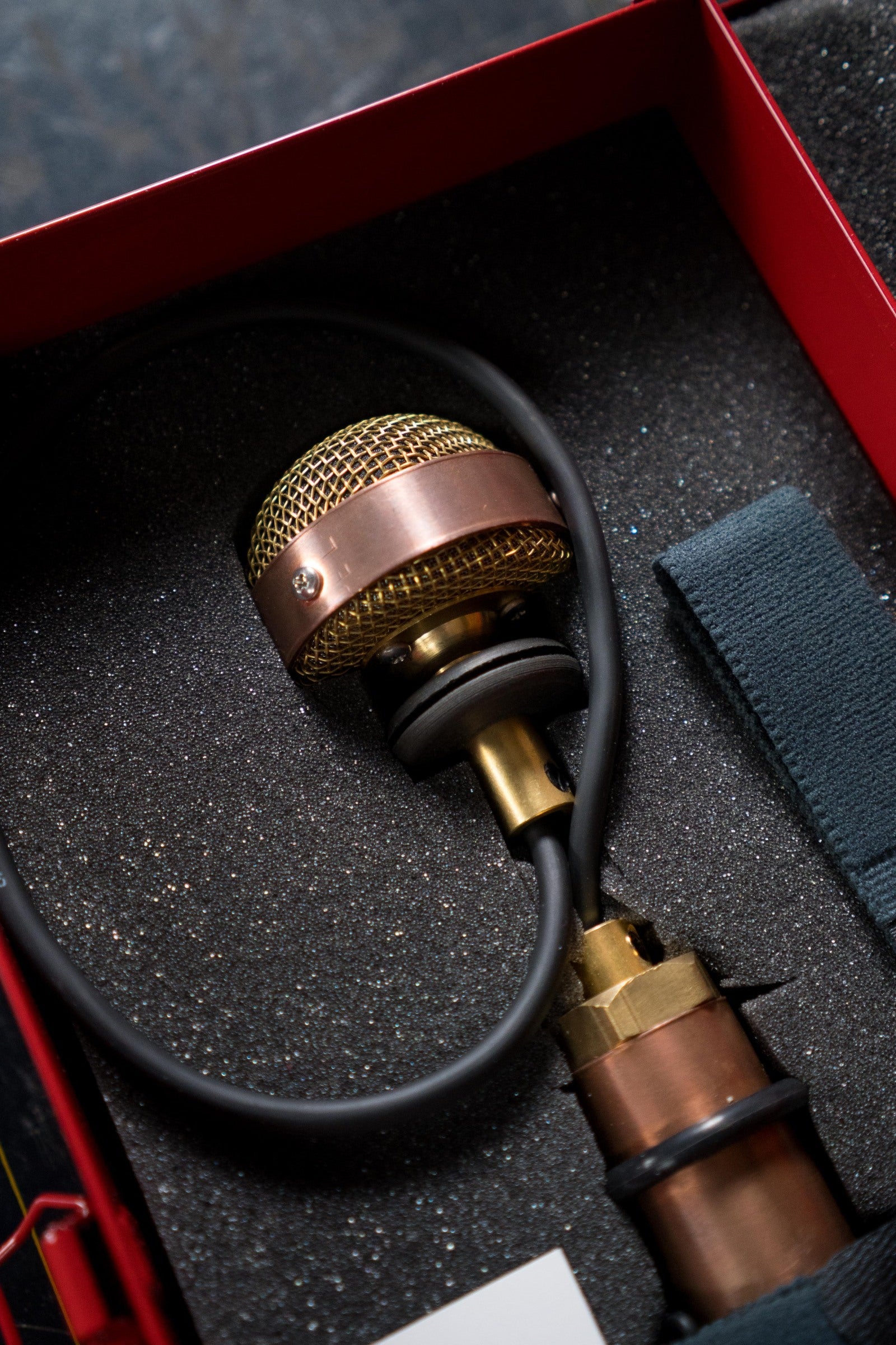 A close-up of the Ear Trumpet Labs Nadine Upright Bass Microphone, featuring a gold mesh grille in a foam-lined red case; a black cable is attached and a strap is visible on the right, delivering natural sound.