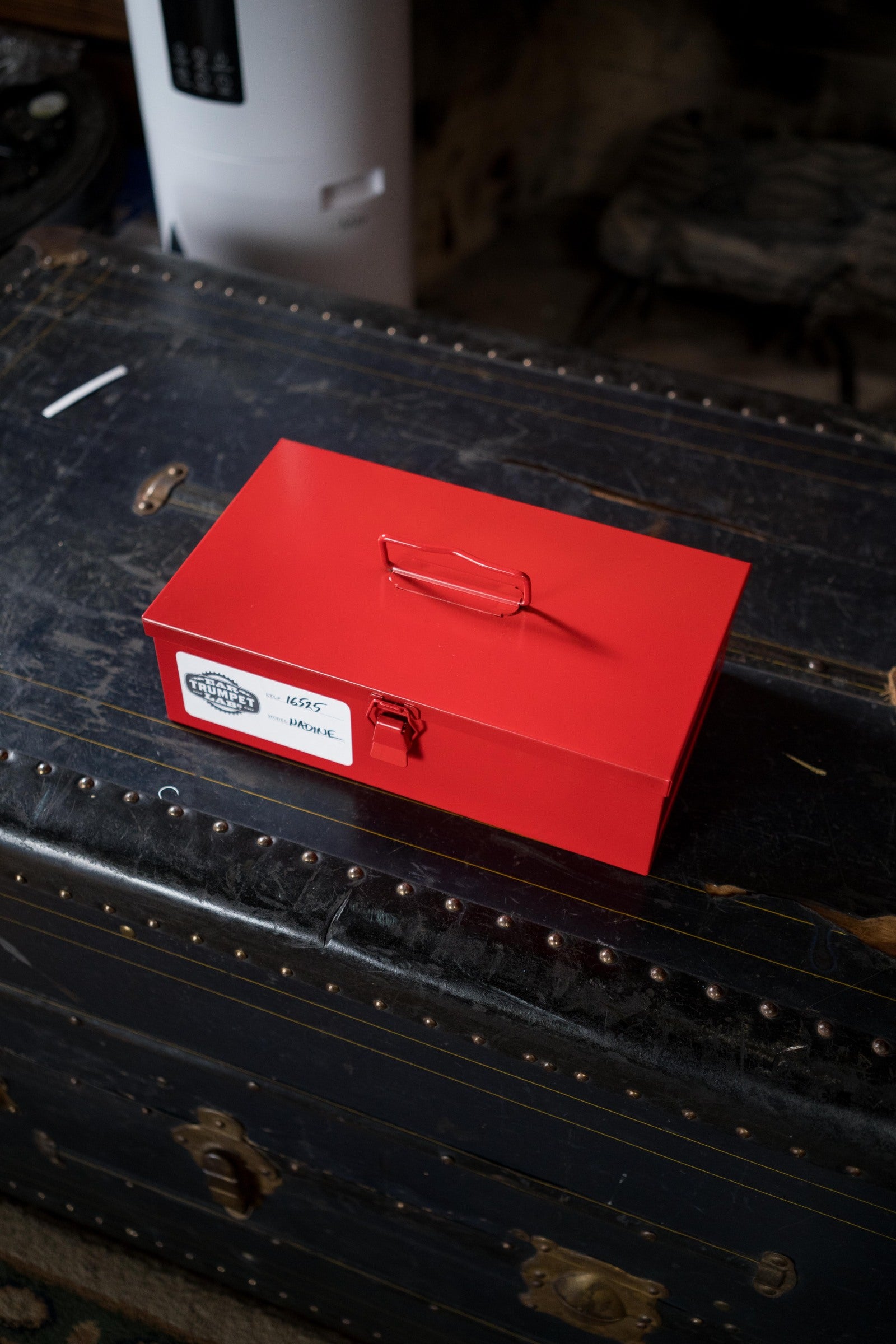 A closed red metal box with a handle sits atop a weathered black trunk labeled KIDS IMAX, while nearby rests an Ear Trumpet Labs Nadine Upright Bass Microphone designed for capturing natural bass sound.