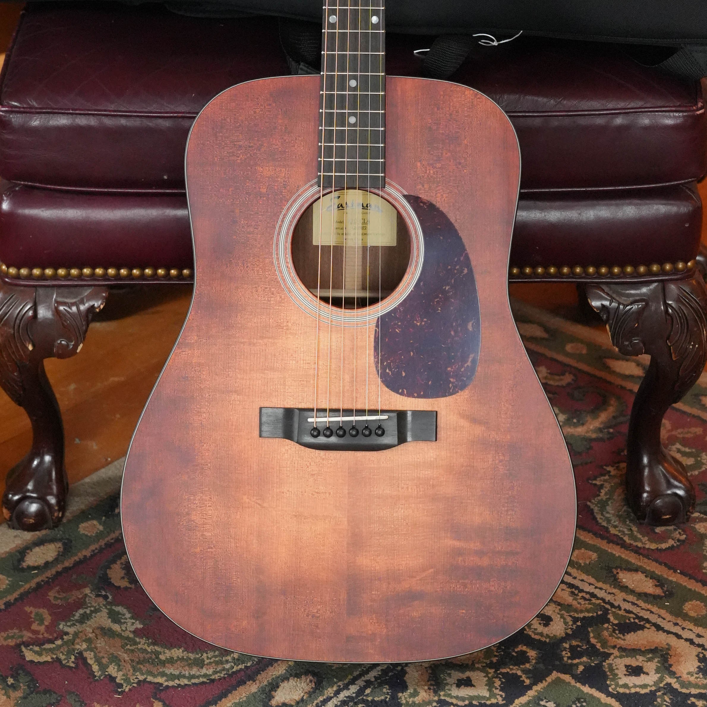 An Eastman Guitars E1D-CLA Classic Finish Dreadnought Acoustic Guitar #1172 with a solid Sitka spruce top stands upright on a patterned rug before a red leather bench with carved wooden legs.