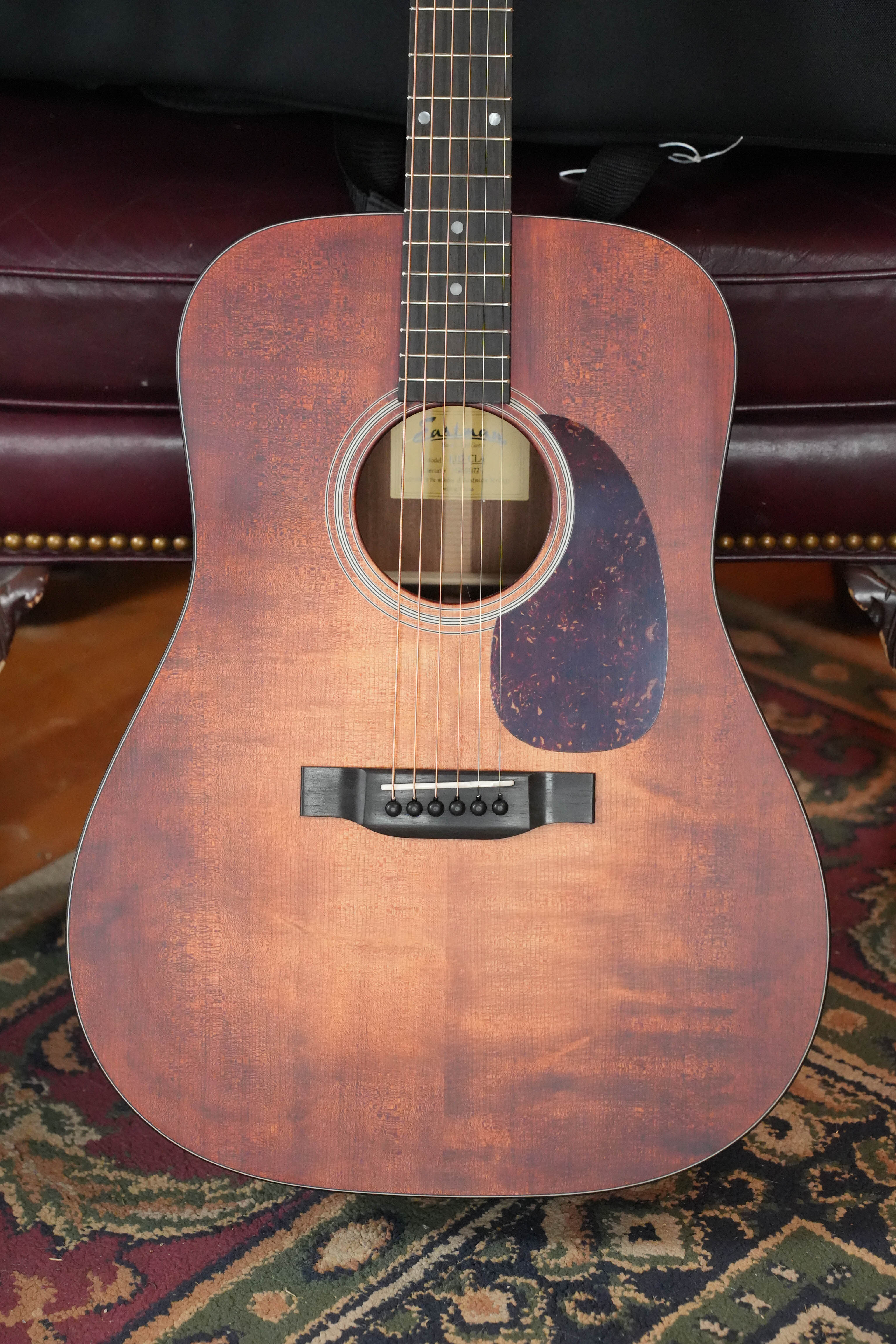 A close-up of an Eastman Guitars E1D-CLA Classic Finish Dreadnought Acoustic Guitar #1172 with a solid Sitka spruce top, resting on a patterned rug in front of a maroon leather chair with brass nailhead trim.