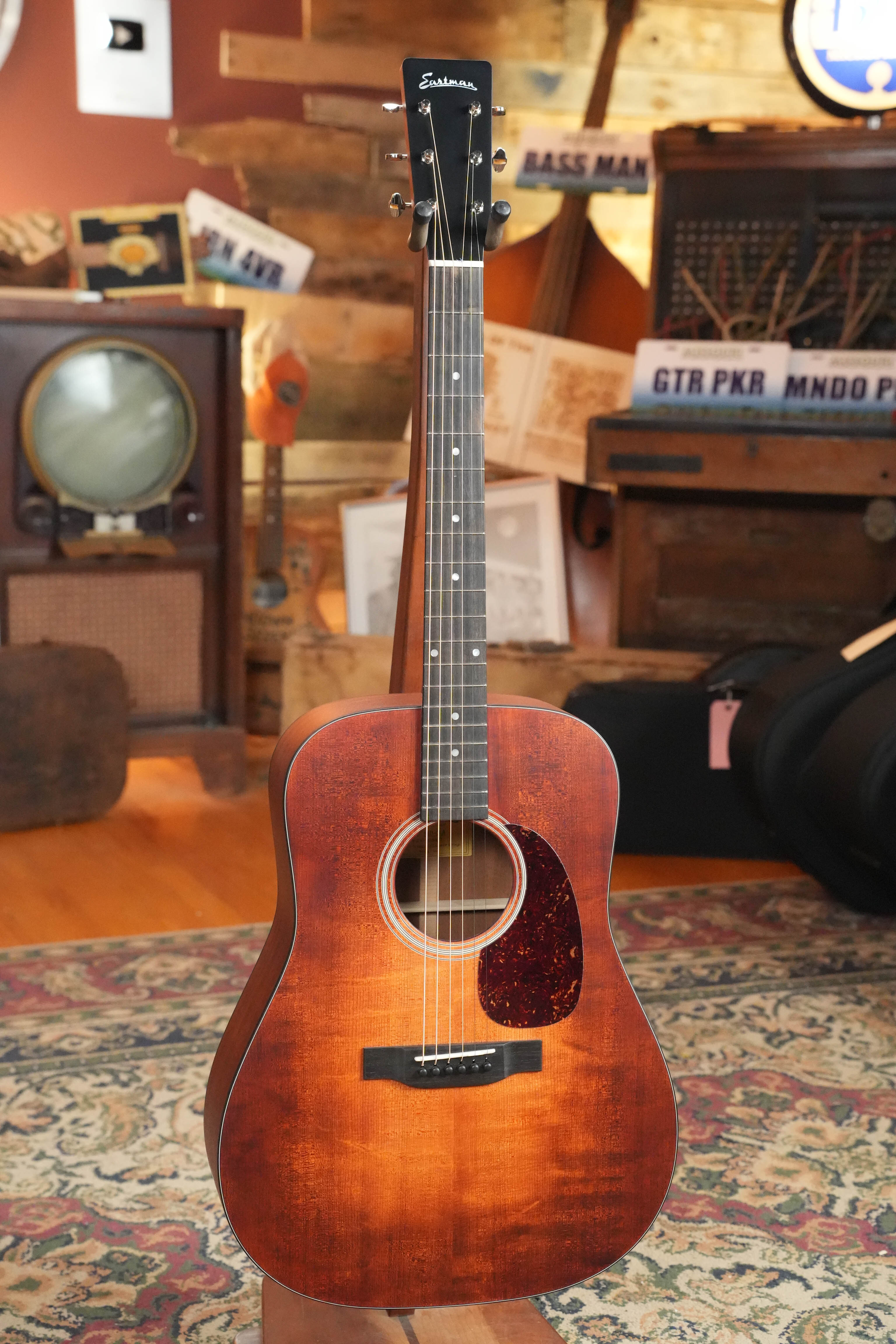An Eastman Guitars E1D-CLA Classic Finish Dreadnought Acoustic Guitar #1172 with a solid Sitka spruce top stands on a patterned rug in a cozy room, surrounded by vintage furniture, musical gear, and wooden walls.
