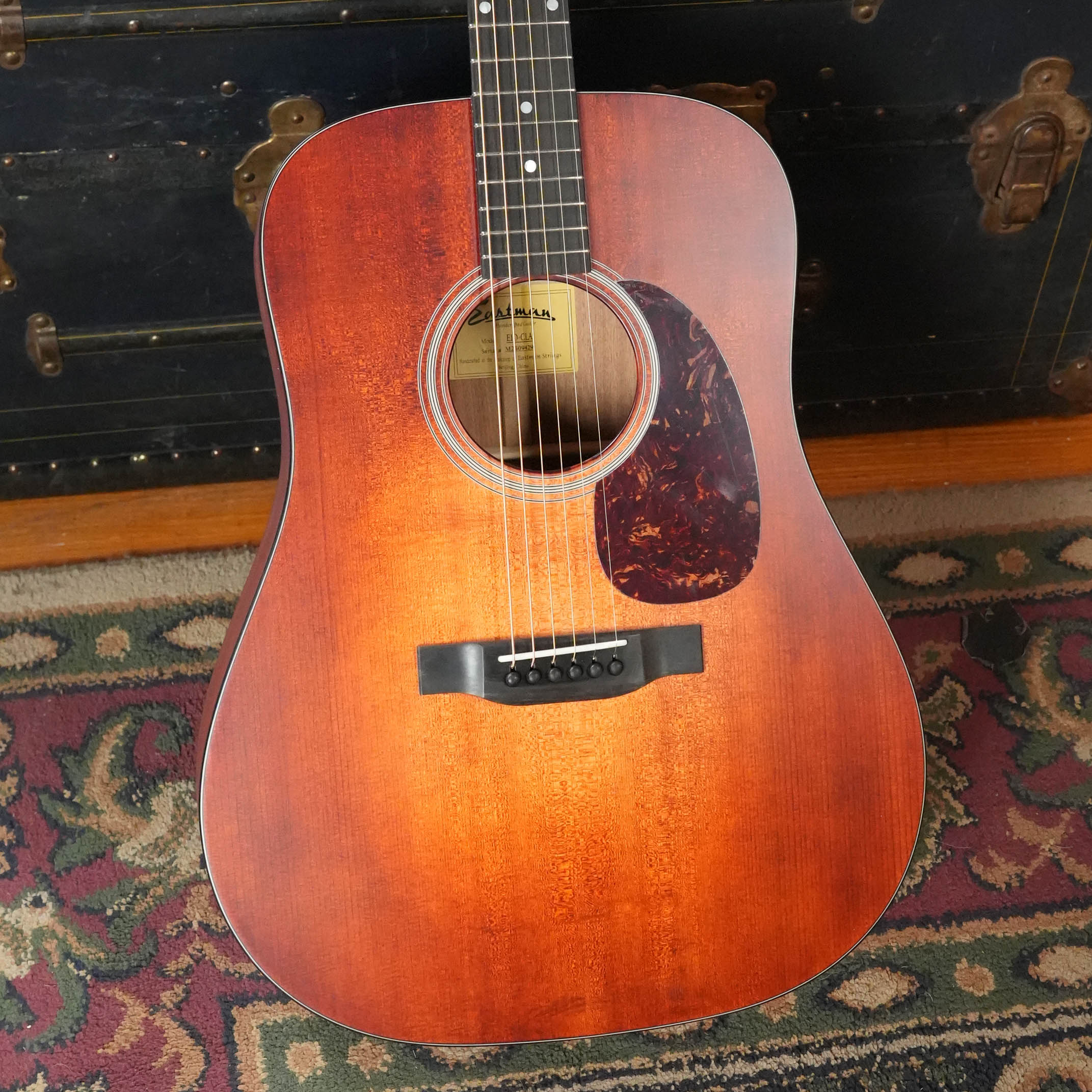 The Eastman Guitars E1D-CLA Classic Finish Dreadnought Acoustic Guitar #9429 stands upright on a patterned rug before a vintage black trunk, its solid Sitka spruce top promising rich tone and clarity.