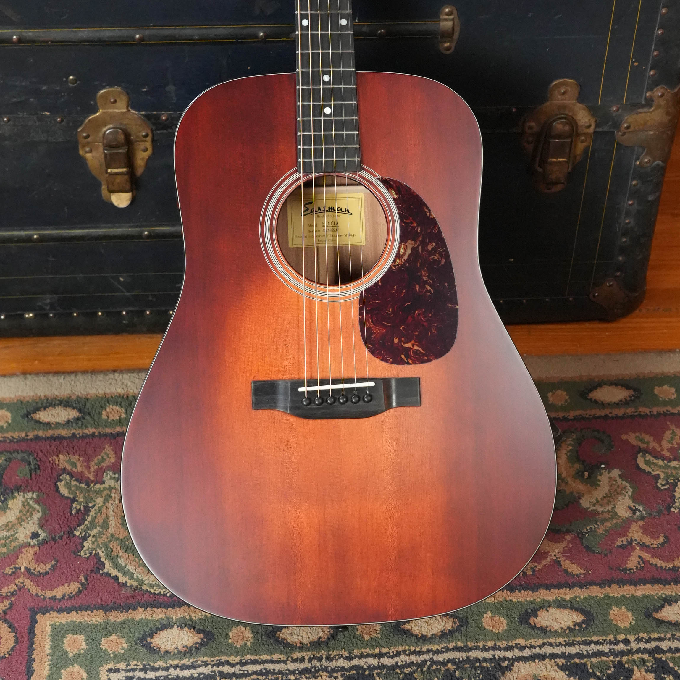The Eastman Guitars E1D-CLA Classic Finish Dreadnought Acoustic Guitar #9747 with a tortoiseshell pickguard rests upright against a dark vintage trunk on a patterned floral rug.