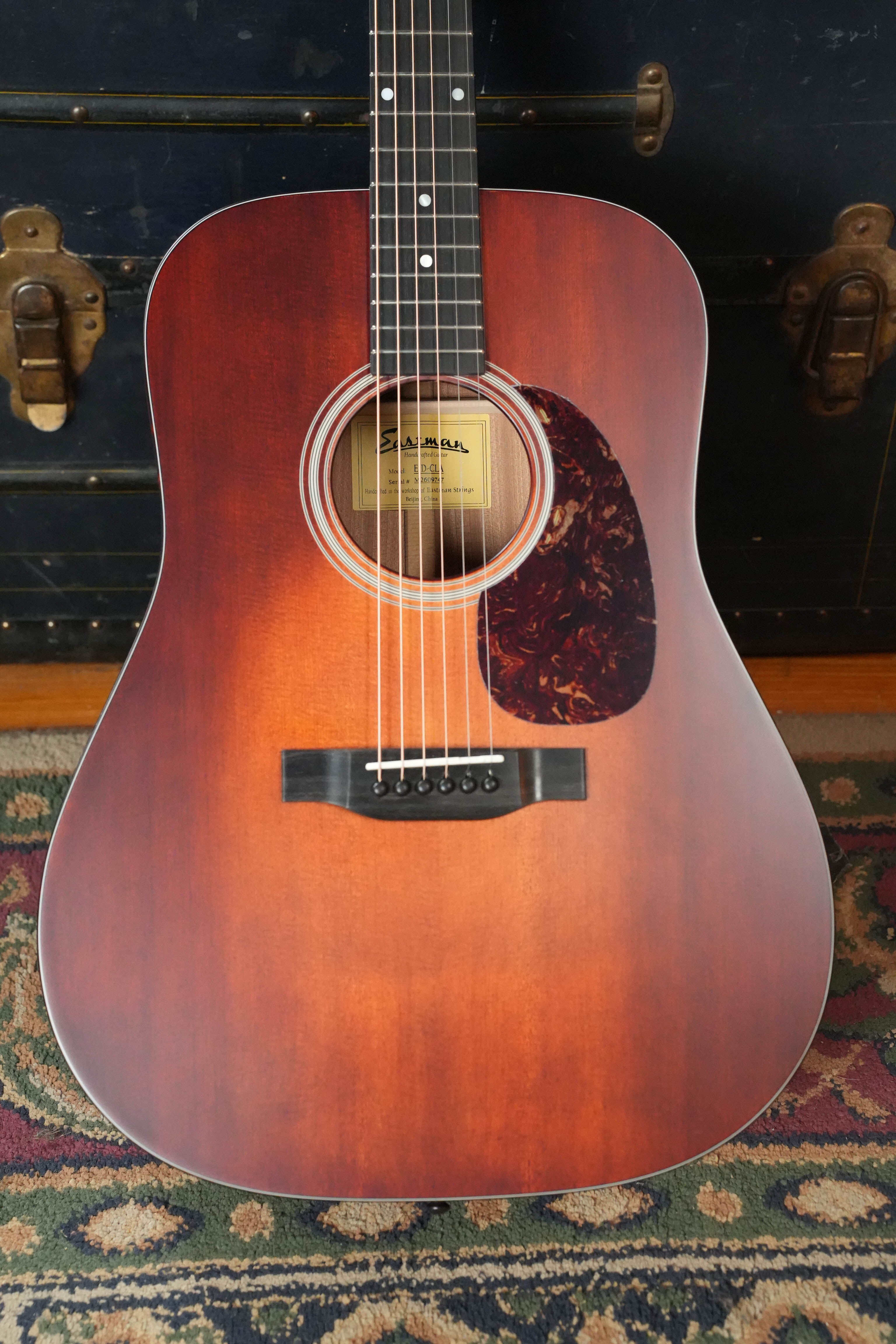 An Eastman E1D-CLA Classic Finish Dreadnought Acoustic Guitar by Eastman Guitars, featuring a tortoiseshell pickguard and solid Sitka spruce top, rests on a patterned rug before a black trunk with brass hardware.