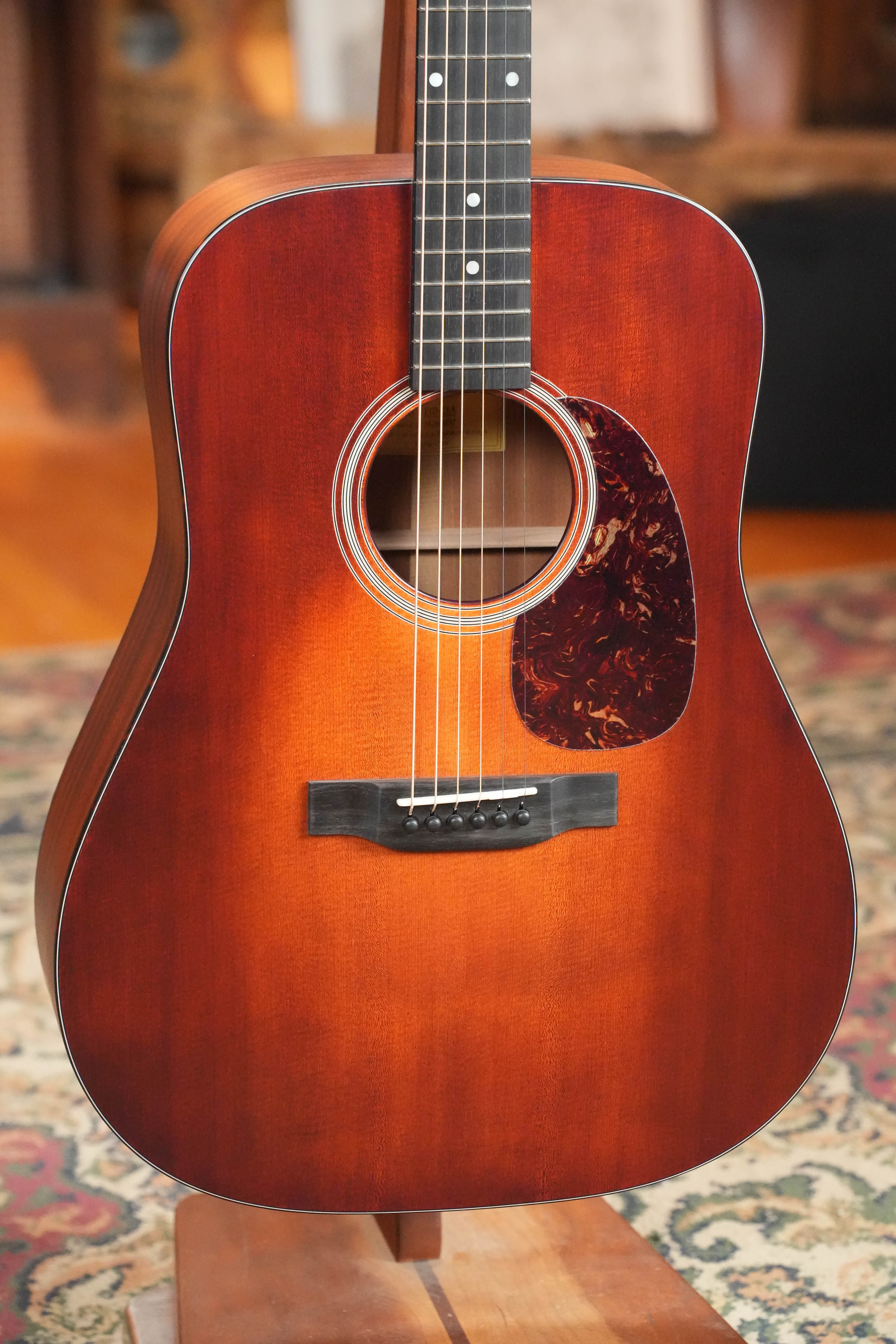 A close-up of the Eastman Guitars E1D-CLA Classic Finish Dreadnought Acoustic Guitar #9747 shows its sunburst top, visible strings, dot inlays, tortoiseshell pickguard, and solid Sitka spruce top on a stand over a patterned rug.