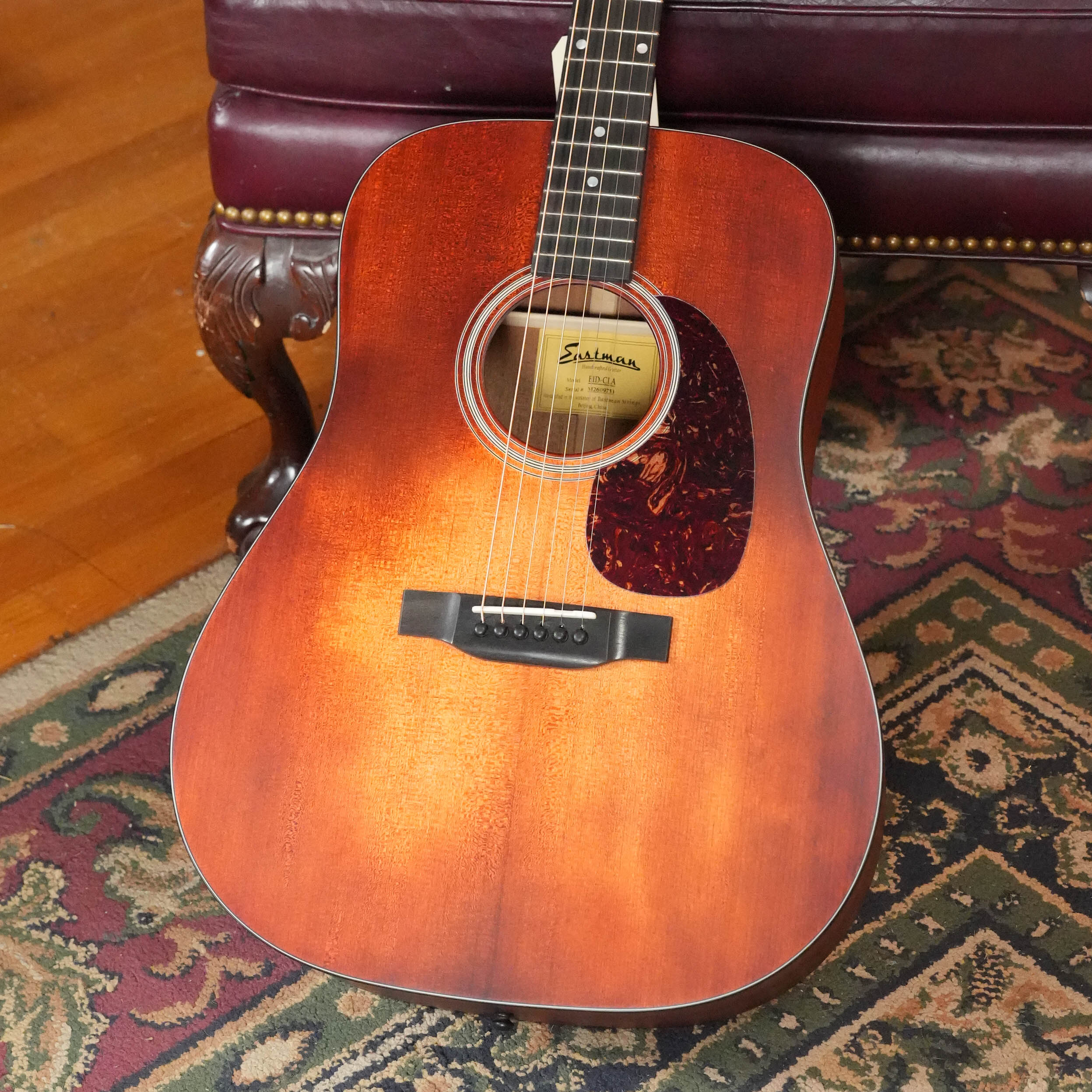 An Eastman E1D-CLA Classic Finish Dreadnought Acoustic Guitar by Eastman Guitars (#9753) rests on a patterned rug in front of a burgundy leather sofa with wood and brass accents.