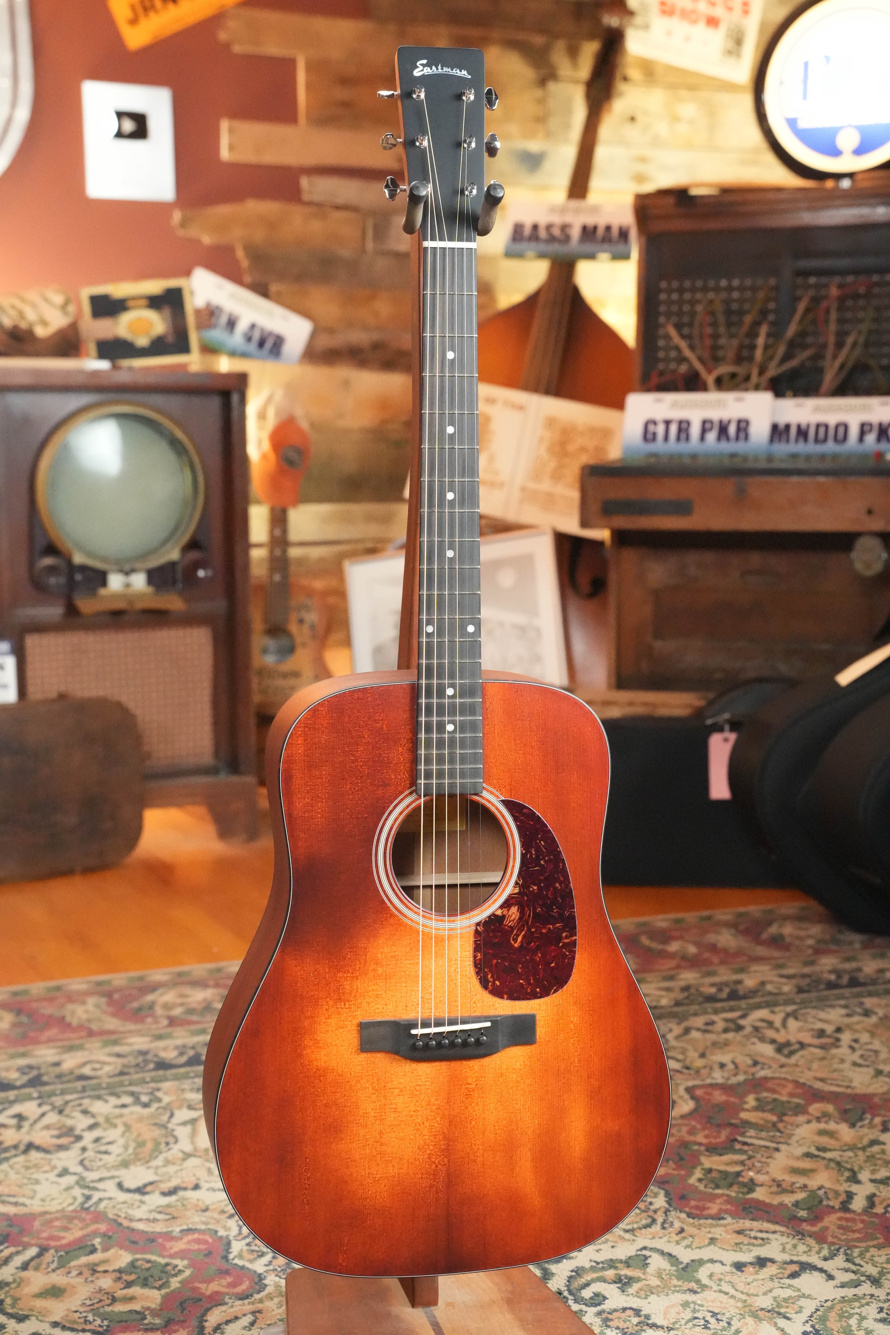 The Eastman Guitars E1D-CLA Classic Finish Dreadnought Acoustic Guitar #9753 stands upright on a patterned rug in a cozy room decorated with retro electronics, musical instruments, and vintage signs.