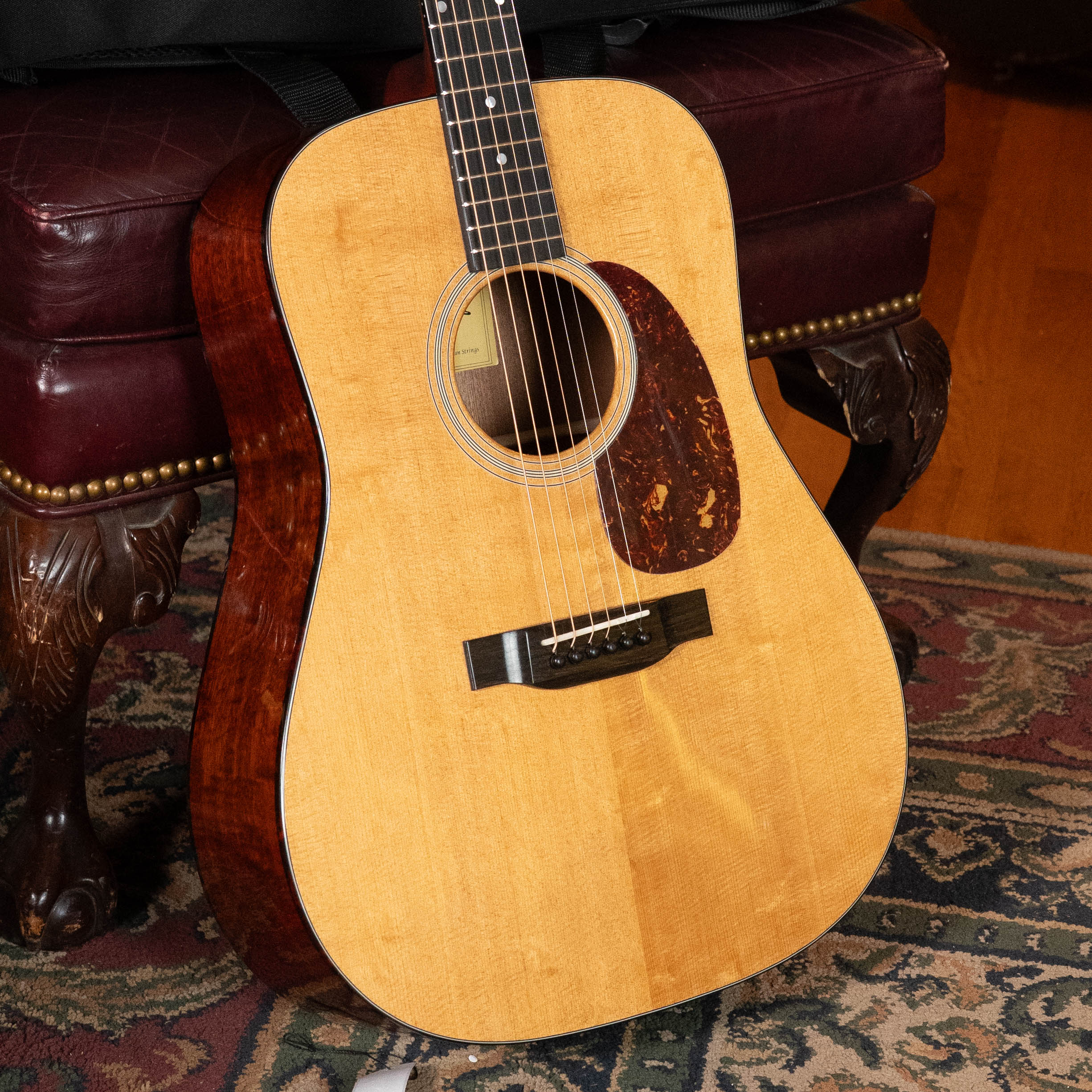 A close-up of the Eastman Guitars E1D-DLX Sitka/Sapele Dreadnought Guitar w/Fishman Pickup #5656, featuring a light wood top and dark sides, resting on a burgundy leather chair beside a multicolored patterned rug.