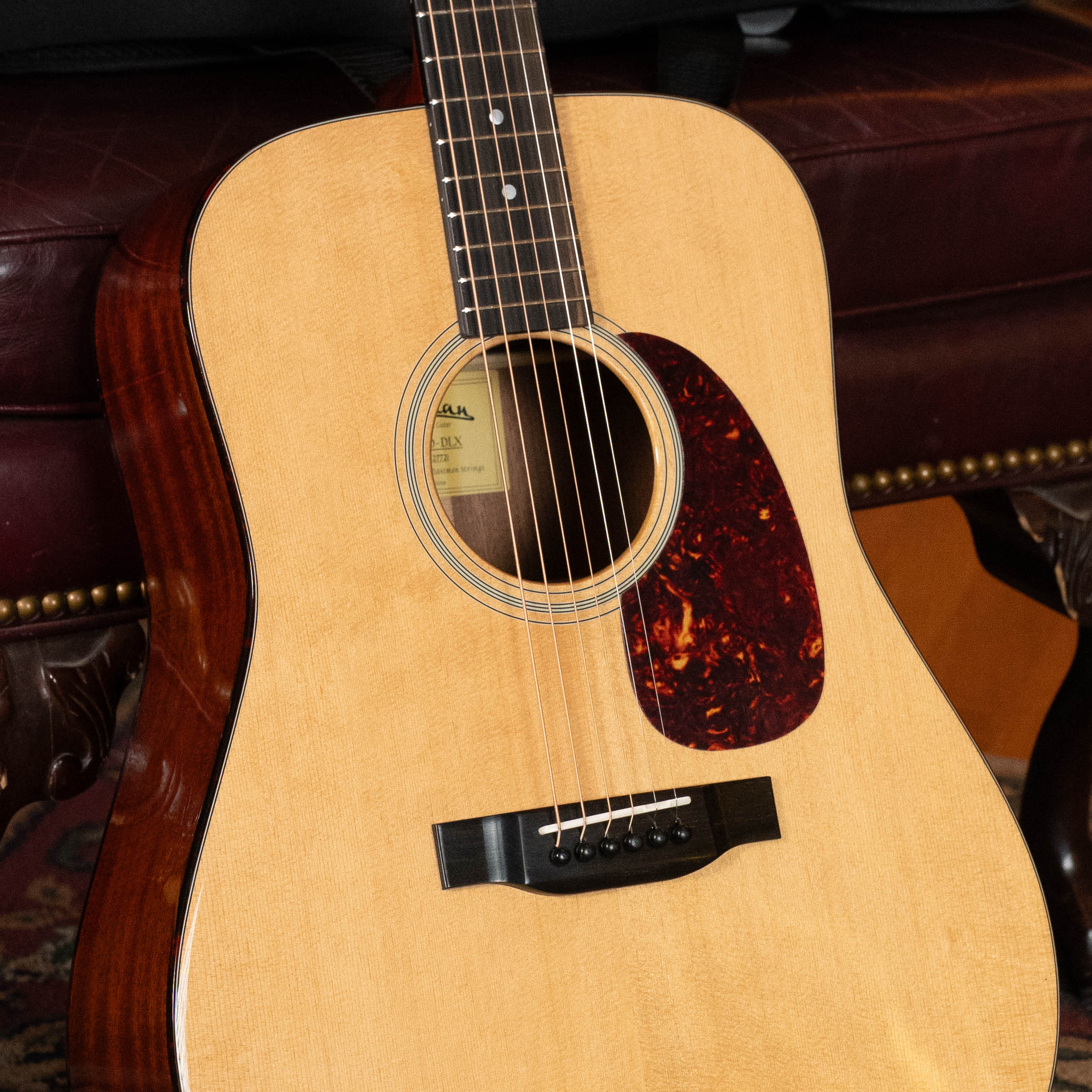A close-up of an Eastman Guitars E1D-DLX Sitka/Sapele Dreadnought Guitar w/Fishman Pickup (#7721), featuring a solid Sitka spruce top and dark pickguard, rests against a maroon leather bench with ornate wooden legs.