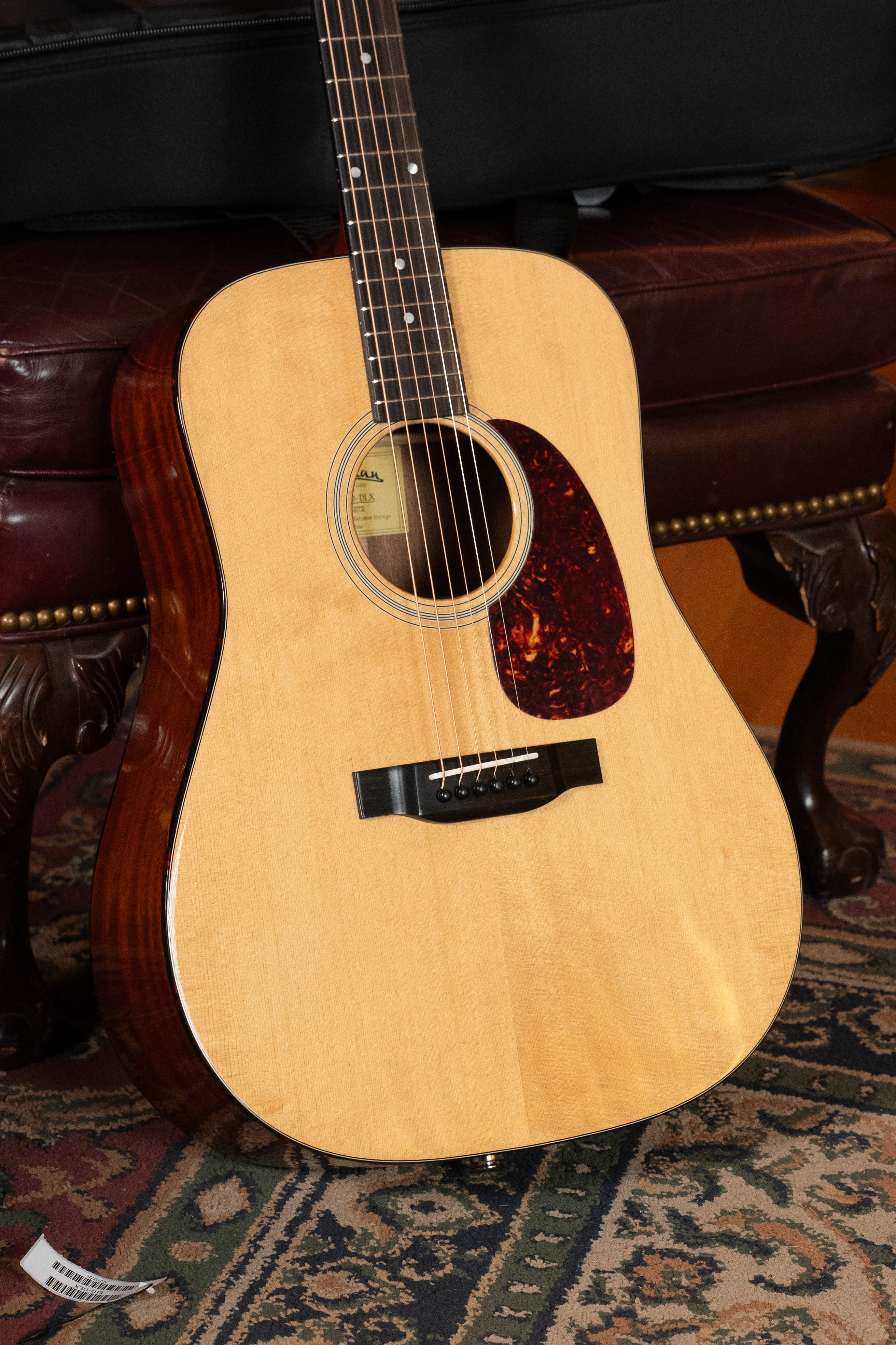 An Eastman Guitars E1D-DLX Sitka/Sapele Dreadnought w/Fishman Pickup (#7721), with a light solid Sitka spruce top and dark pickguard, leans against a maroon leather chair on a patterned rug; part of its case is visible.