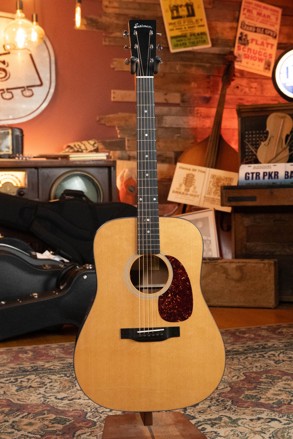 An Eastman Guitars E1D-DLX Sitka/Sapele Dreadnought w/Fishman Pickup #8436 with a light wood finish and dark pickguard stands upright in a cozy, vintage-style room filled with instruments and posters.