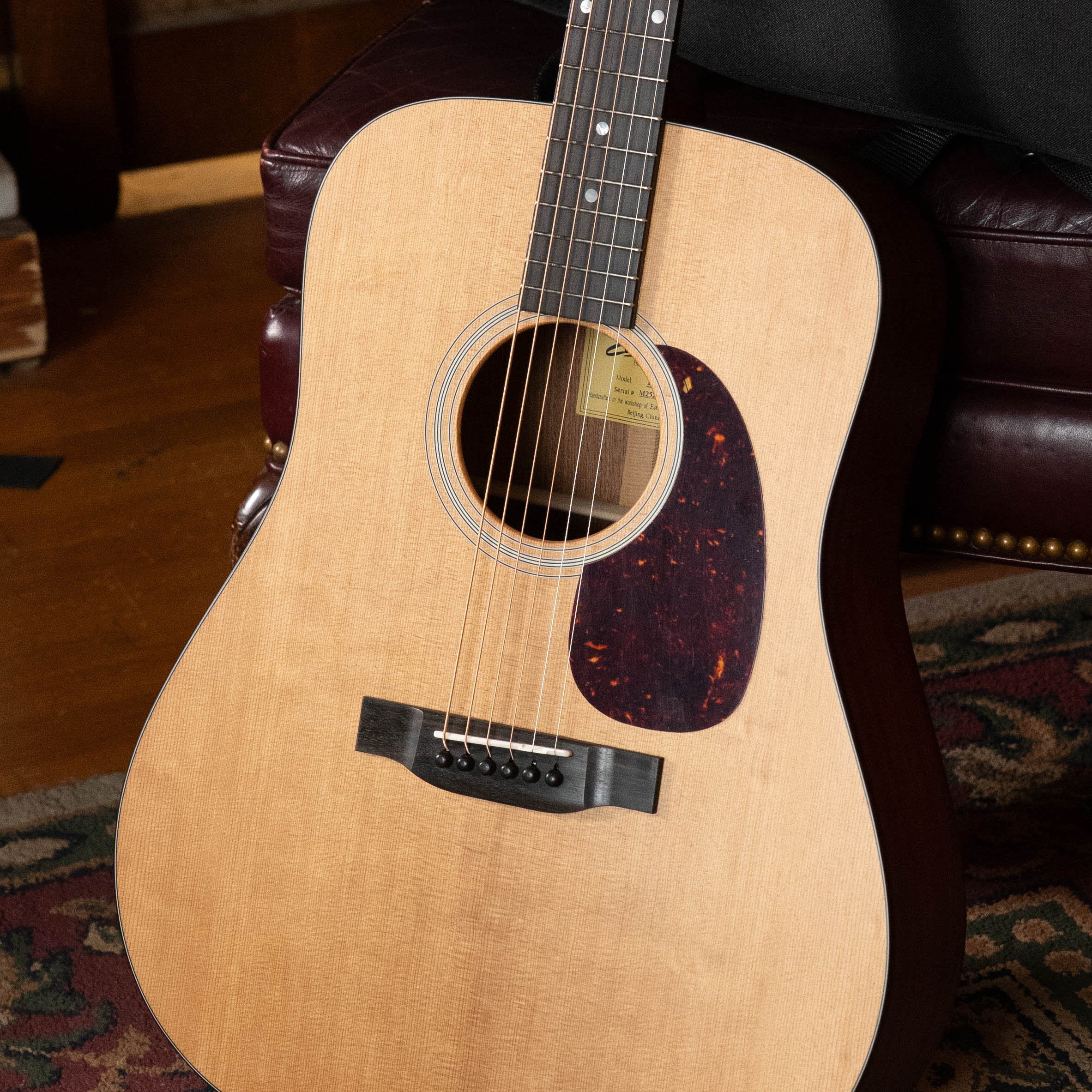 A close-up of an Eastman Guitars E1D Sitka/Sapele Dreadnought Acoustic Guitar #6855, featuring a solid Sitka spruce top, on a leather chair. Its body, strings, and pickguard are visible with a patterned rug and wooden floor behind.