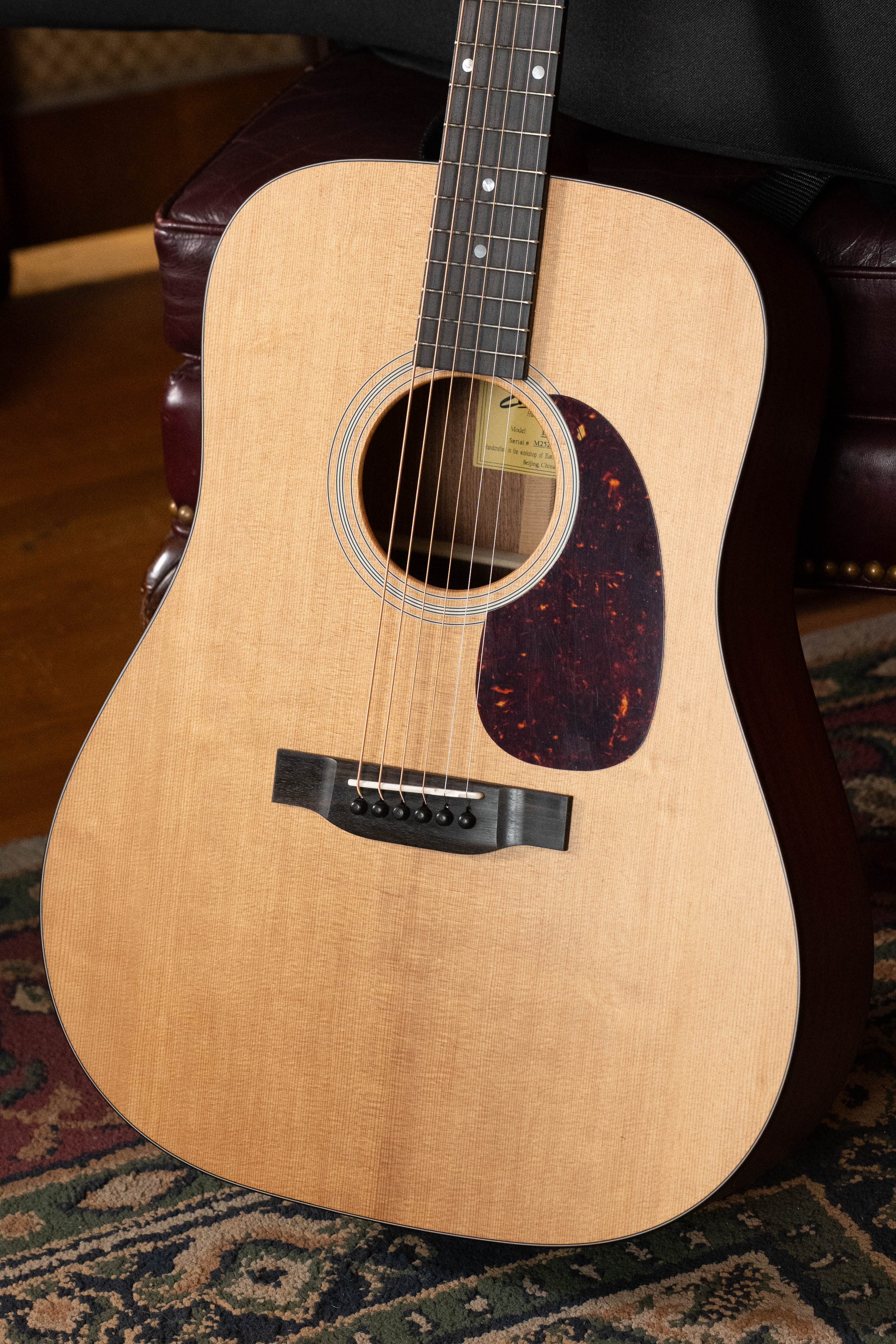 A close-up of the Eastman Guitars E1D Sitka/Sapele Dreadnought Acoustic Guitar #6855 featuring a solid Sitka spruce top, dark pickguard, and six strings, resting on a patterned rug beside a leather chair.