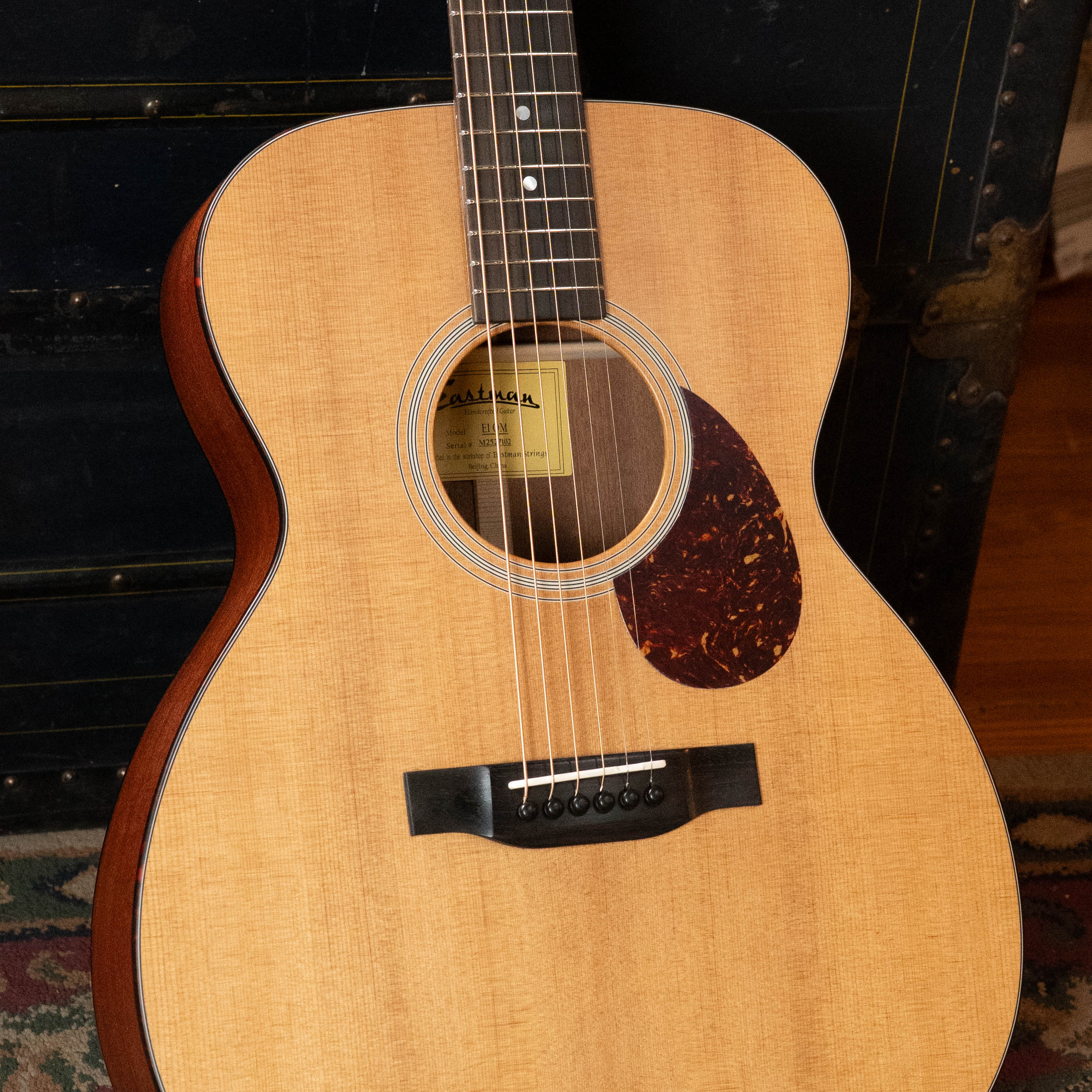 An Eastman Guitars E1OM All Solid Sitka/Sapele Orchestra Model Acoustic Guitar #7102 with six strings and a tortoiseshell pickguard stands upright before a dark trunk, showing its solid Sitka Spruce top and the brand label inside the soundhole.