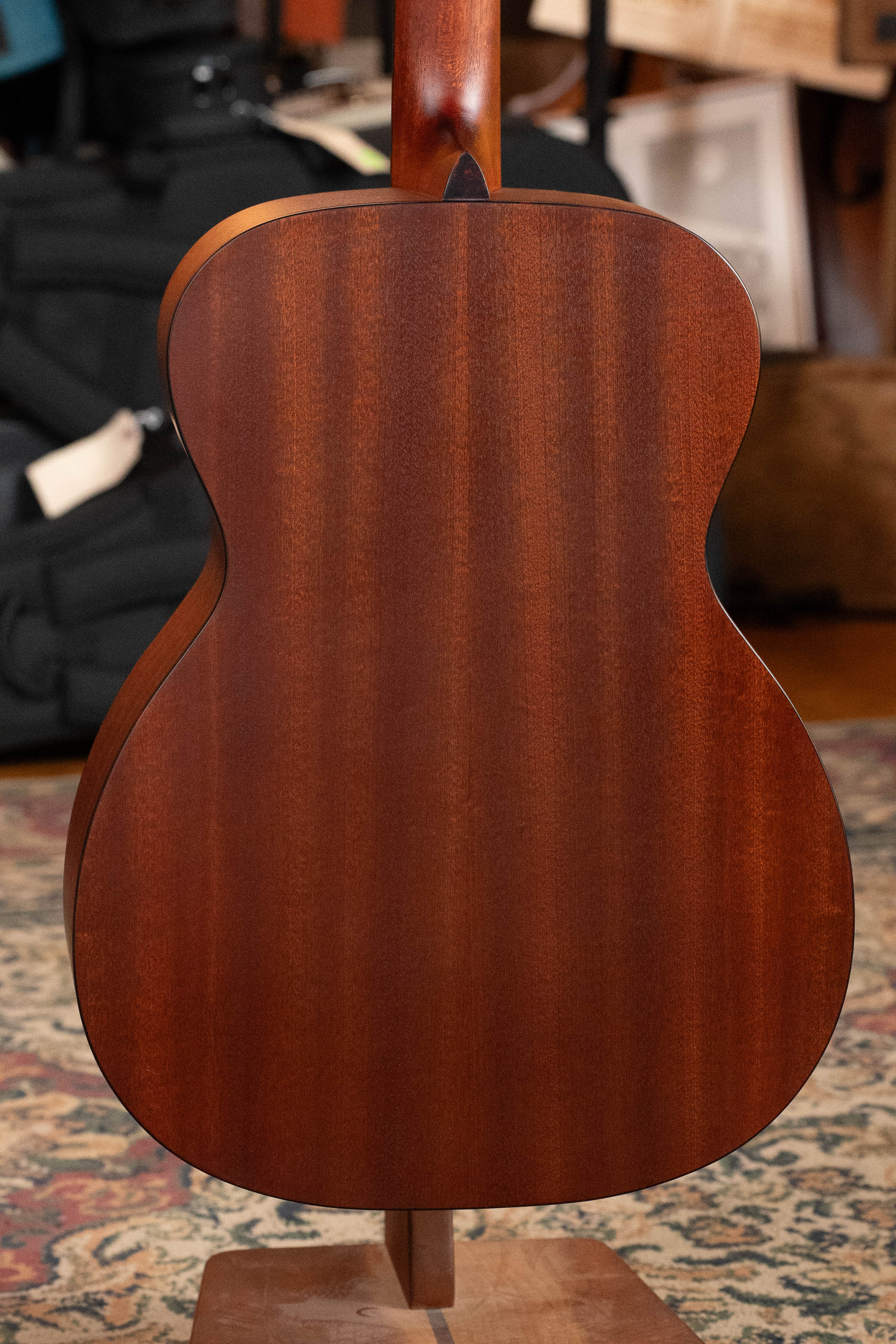 A close-up of the back of an Eastman Guitars E1OM All Solid Sitka/Sapele Orchestra Model Acoustic Guitar #7102 with a rich reddish-brown finish, standing upright on a stand over a patterned rug, blurred musical gear in the background.