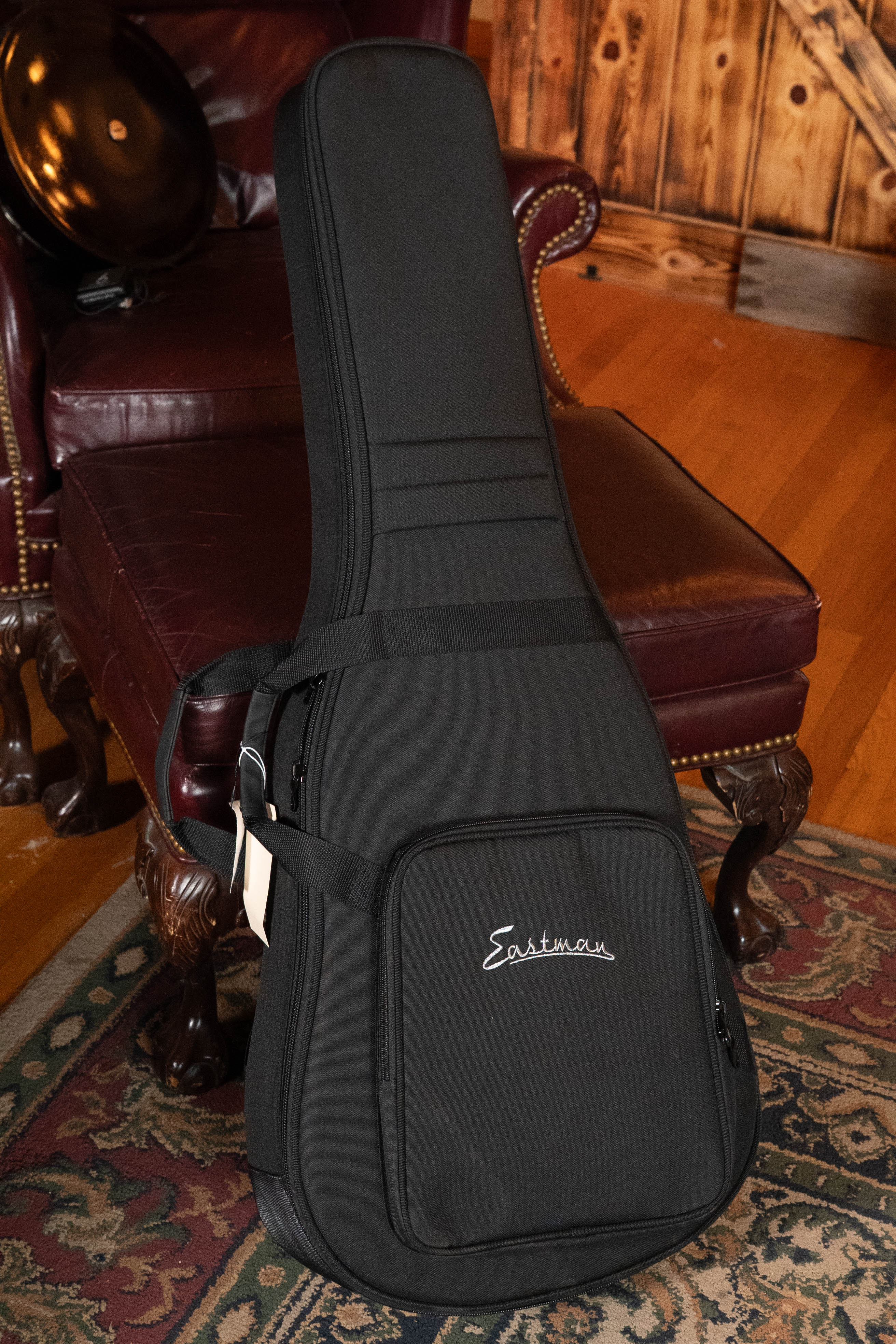 An Eastman Guitars E1OM All Solid Sitka/Sapele Orchestra Model Acoustic Guitar #7102 in a black case stands upright by a maroon leather chair in a room with wooden floors and rustic wood-paneled walls.