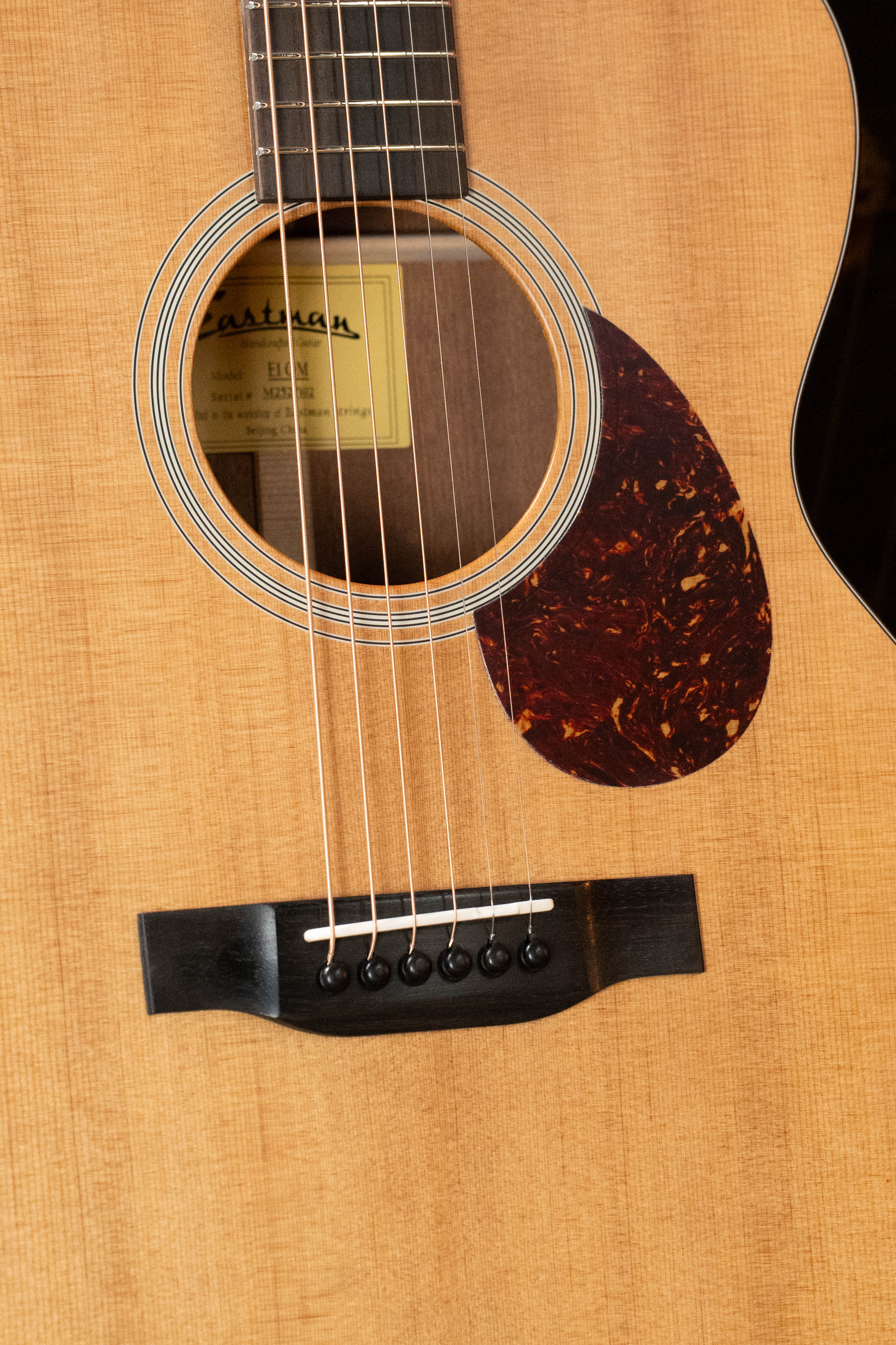 Close-up of an Eastman Guitars E1OM All Solid Sitka/Sapele Orchestra Model Acoustic Guitar #7102, showing the natural Sitka spruce top, tortoiseshell-style pickguard, sound hole, strings, bridge, and part of the label.