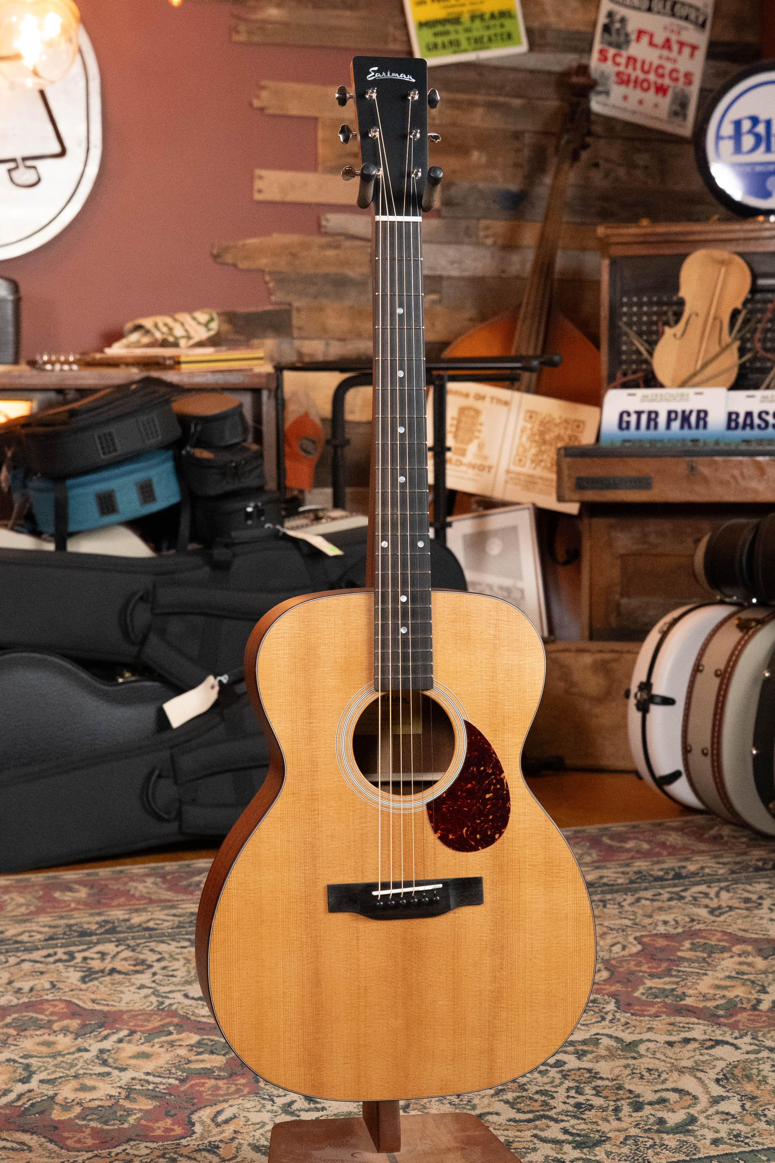 An Eastman Guitars E1OM All Solid Sitka/Sapele Orchestra Model Acoustic Guitar #7102 stands on a patterned rug in a cozy, rustic room filled with musical instruments, cases, and vintage decor.