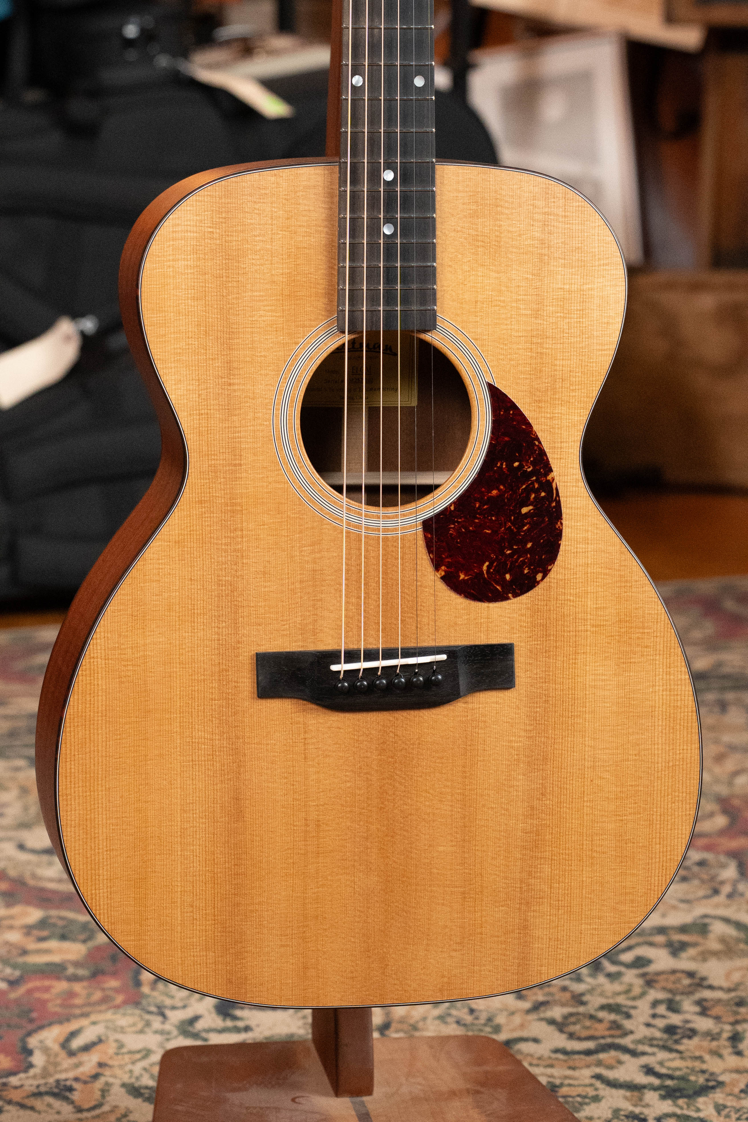 An Eastman Guitars E1OM All Solid Sitka/Sapele Orchestra Model Acoustic Guitar #7102 with a natural finish stands on display, featuring a solid Sitka spruce top, six strings, sound hole, dark pickguard, and a patterned rug background.