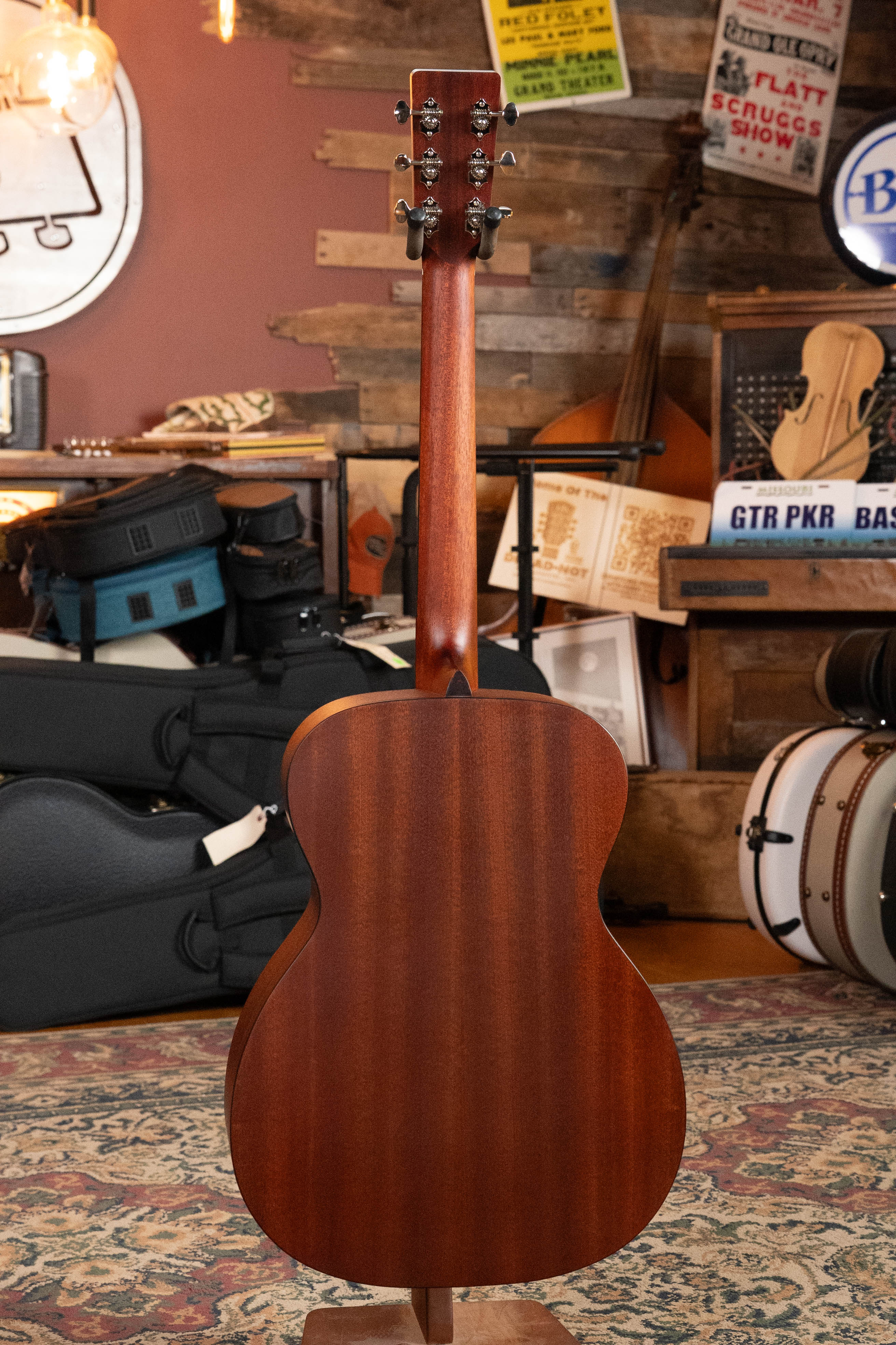 The Eastman E1OM All Solid Sitka/Sapele Orchestra Model Acoustic Guitar #7102 by Eastman Guitars is pictured standing upright in a cozy, rustic room surrounded by musical instruments, guitar cases, drums, and unique wall decor.