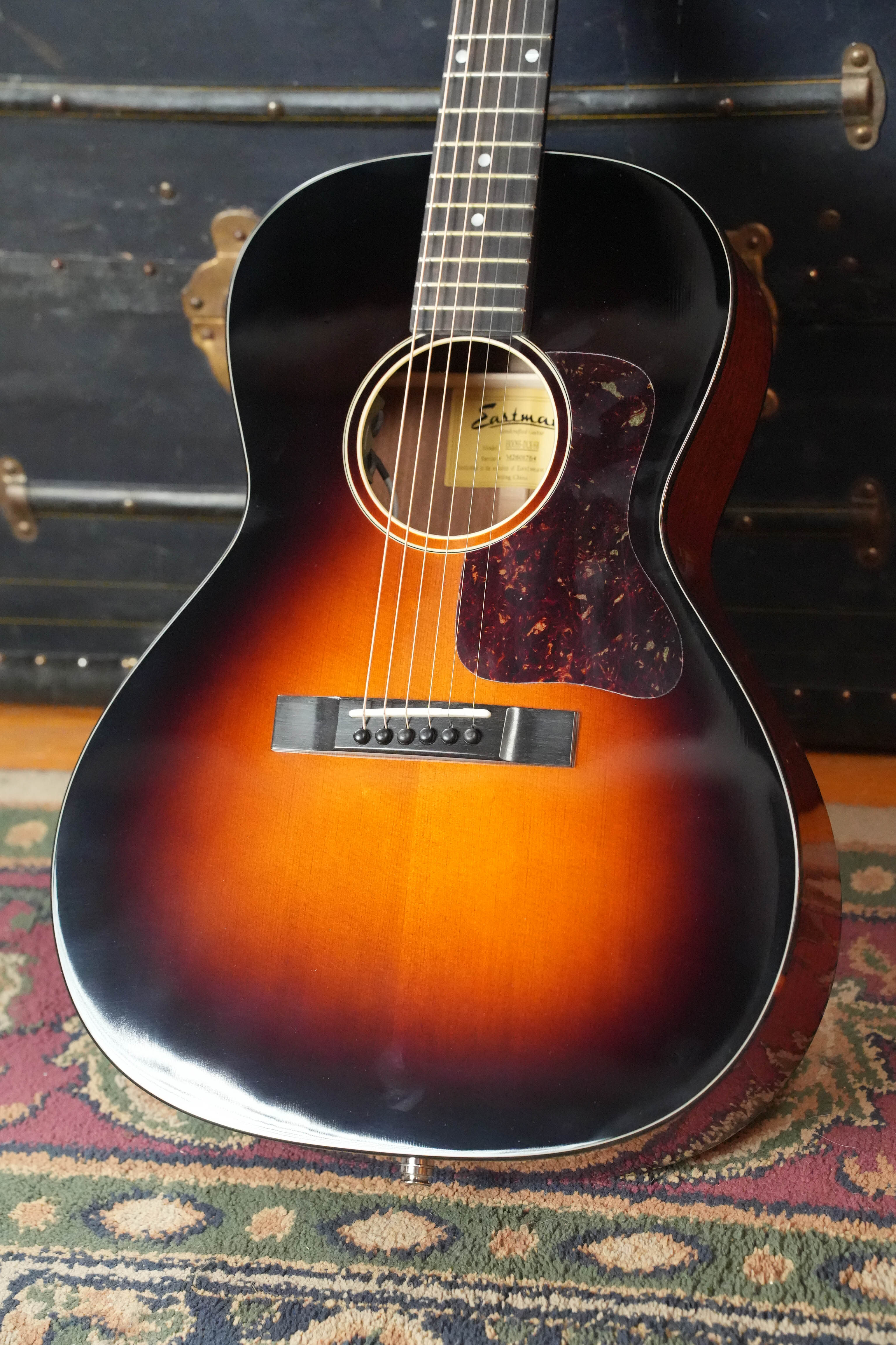 The Eastman Guitars E1OOSS-DLX-SB Sunburst 00 Acoustic-Electric Guitar #1764 stands upright on a patterned rug, its Truetone sunburst finish and tortoiseshell pickguard evoking pre-war charm beside a vintage-style trunk.