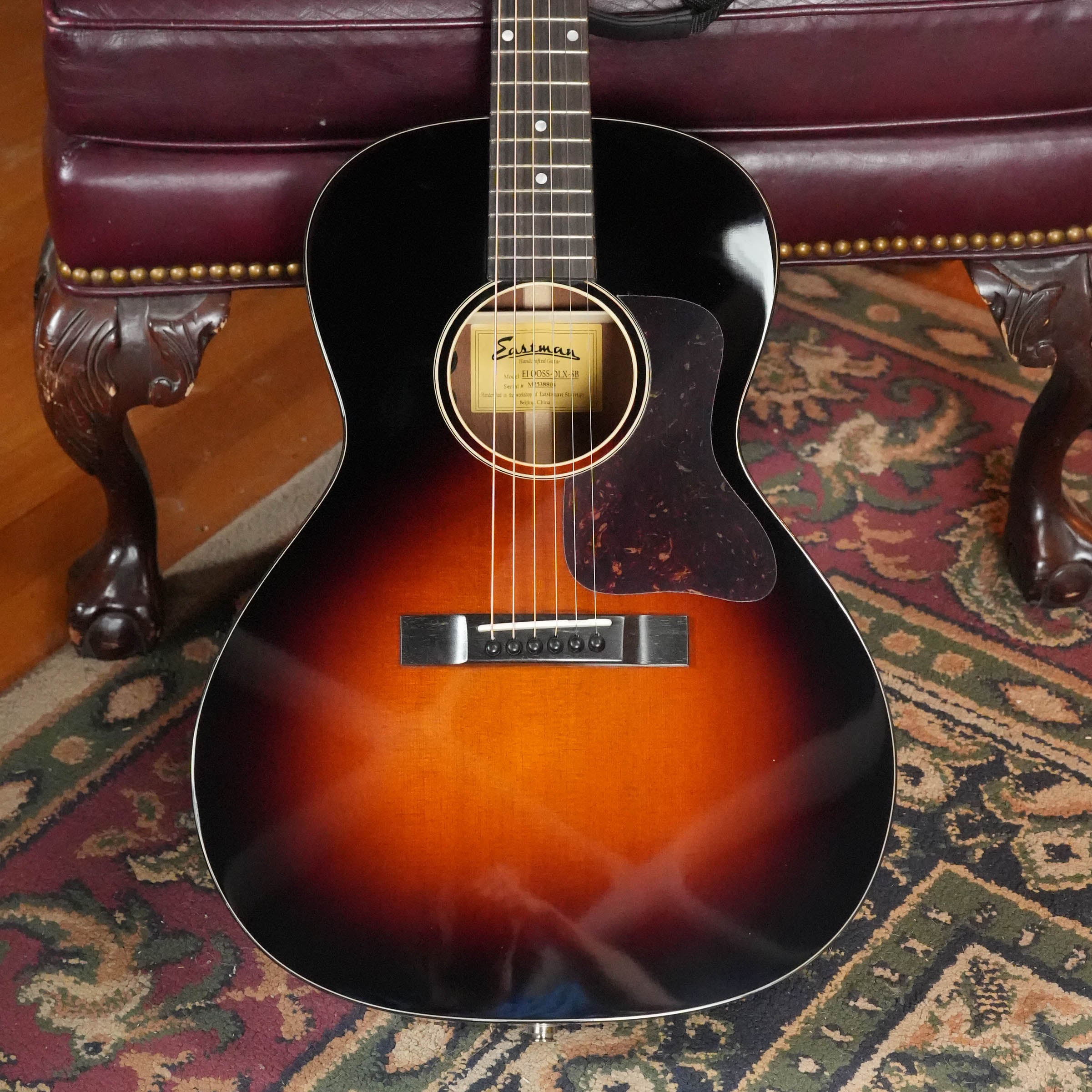 The Eastman Guitars E1OOSS-DLX-SB Sunburst 00 Acoustic-Electric Guitar #8804 leans against a dark, ornate leather chair with brass studs, resting on a floral-patterned rug and radiating a pre-war vibe.