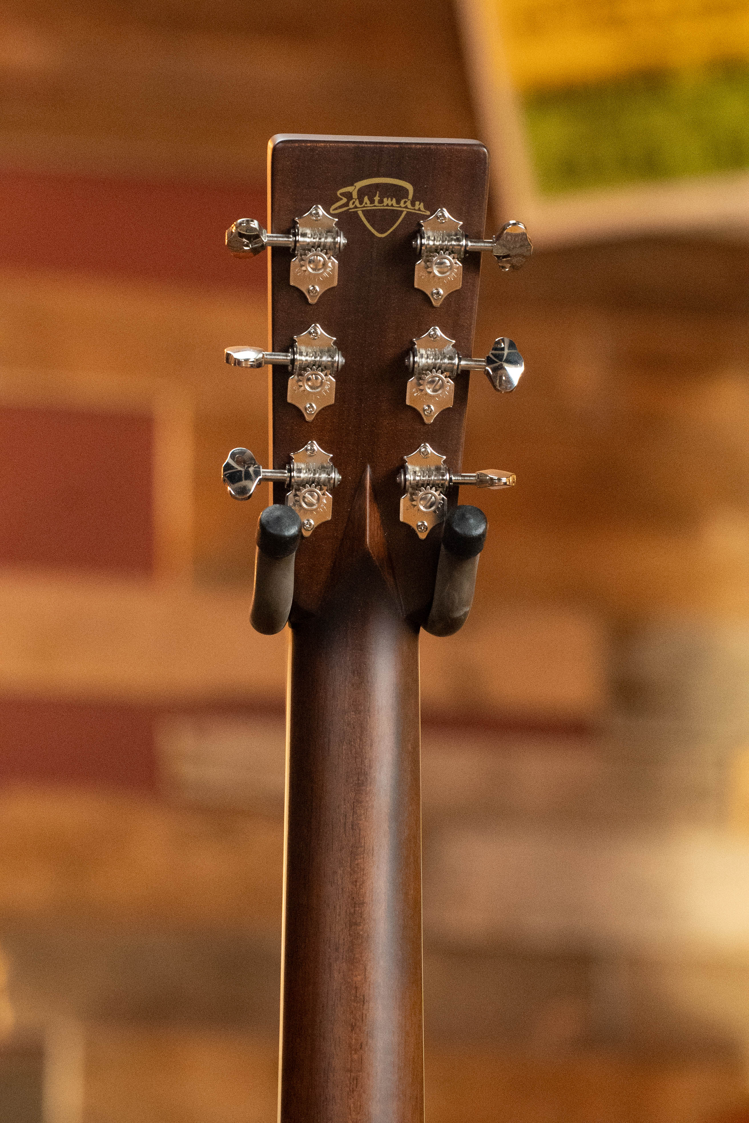 The back of an Eastman E20D-LSH-TC acoustic guitar headstock with six silver tuners and the Eastman Guitars logo rests on a wall-mounted holder against a blurred wooden background.
