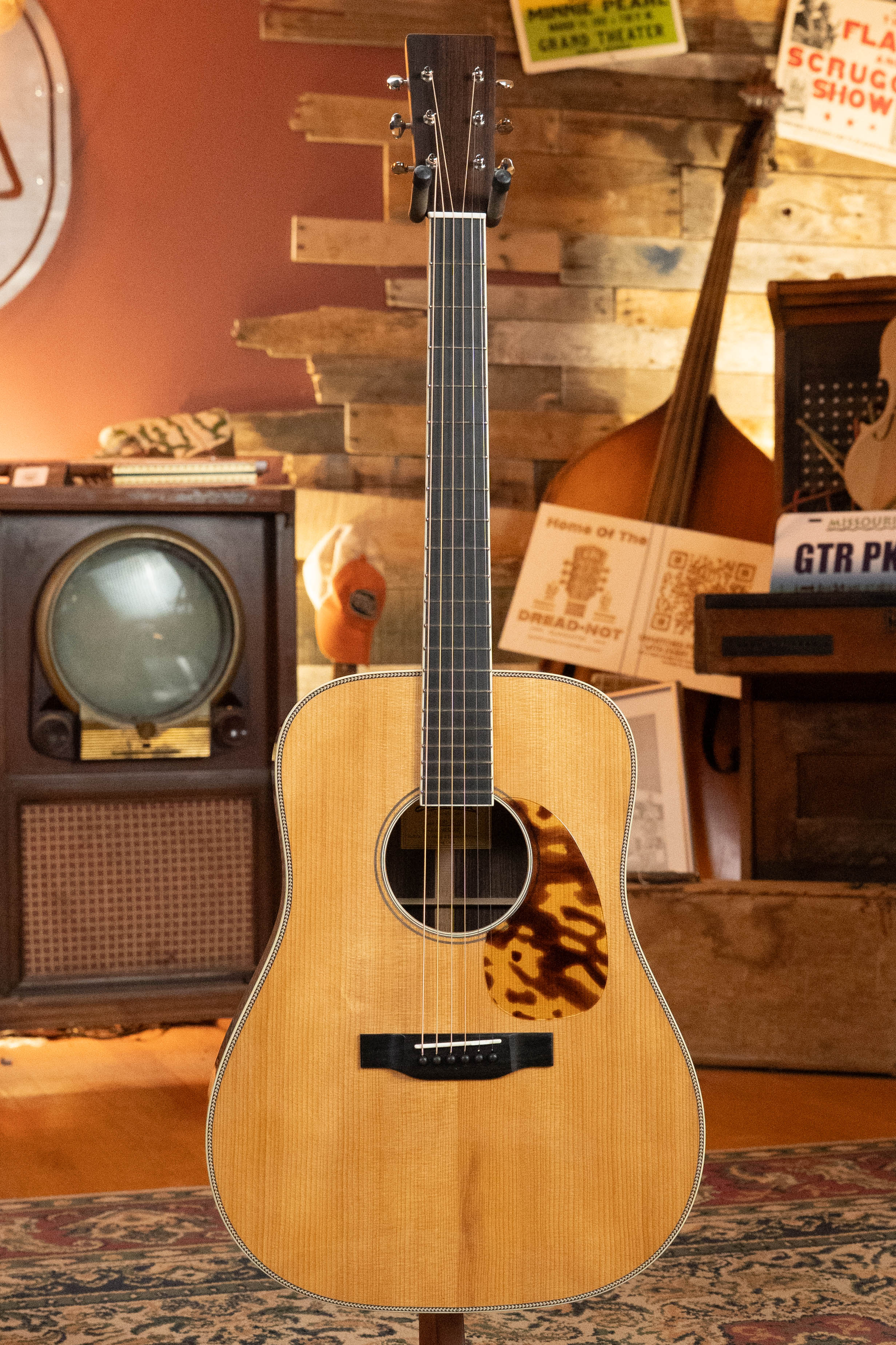 An Eastman Guitars E20D-LSH-TC Thermo-Cured Adirondack/Rosewood Dreadnought #3917 stands upright in a cozy vintage room, surrounded by a retro TV, upright bass, and warm wooden decor.