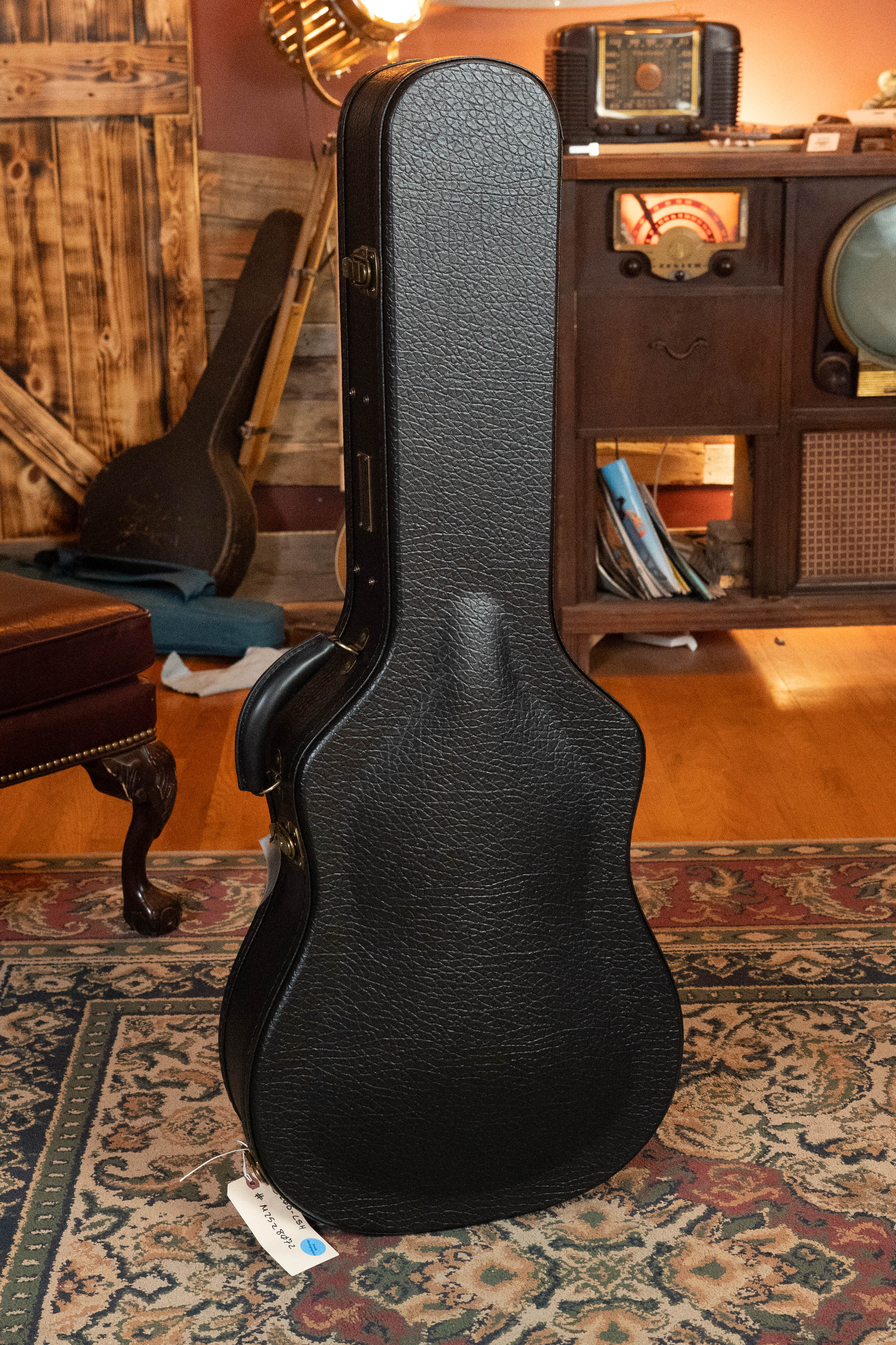 A black acoustic guitar case stands upright on a patterned rug in a cozy room, possibly safeguarding an Eastman Guitars E20D-LSH-TC Thermo-Cured Adirondack/Rosewood Large Soundhole Dreadnought Acoustic Guitar #8072.