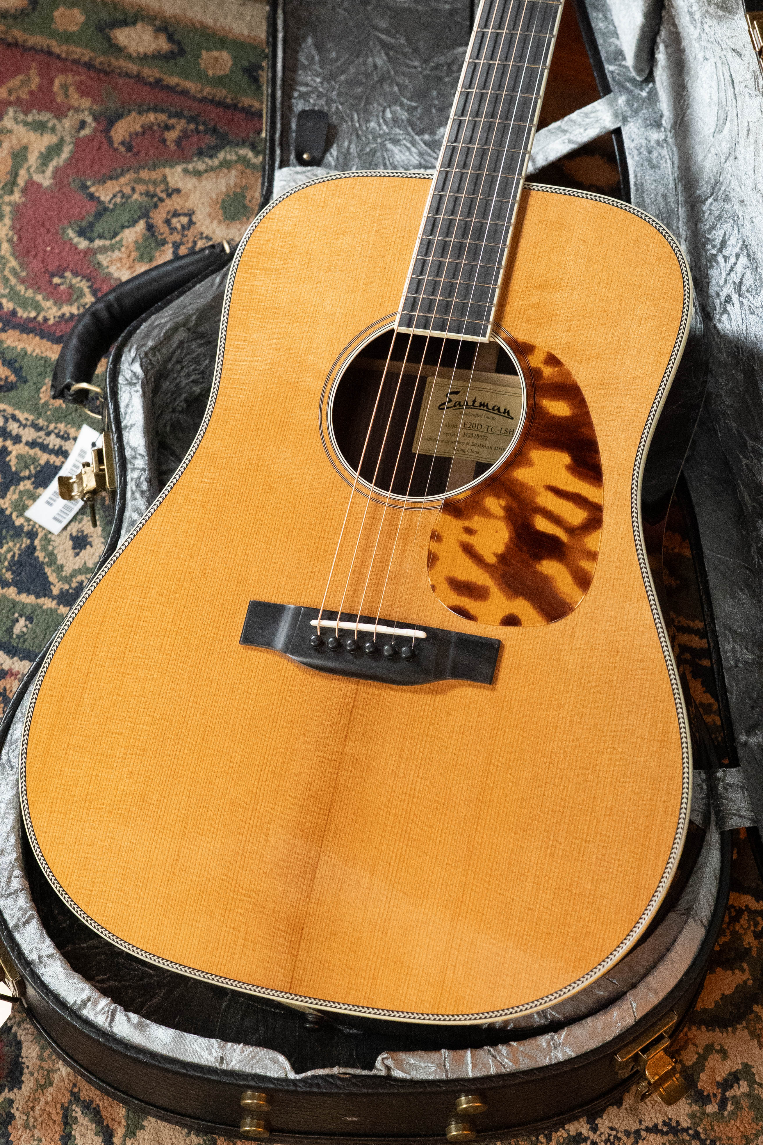 A close-up of an Eastman Guitars E20D-LSH-TC Thermo-Cured Adirondack/Rosewood Large Soundhole Dreadnought Acoustic Guitar #8072 with a tortoiseshell pickguard, resting in an open hard case on a patterned carpet.