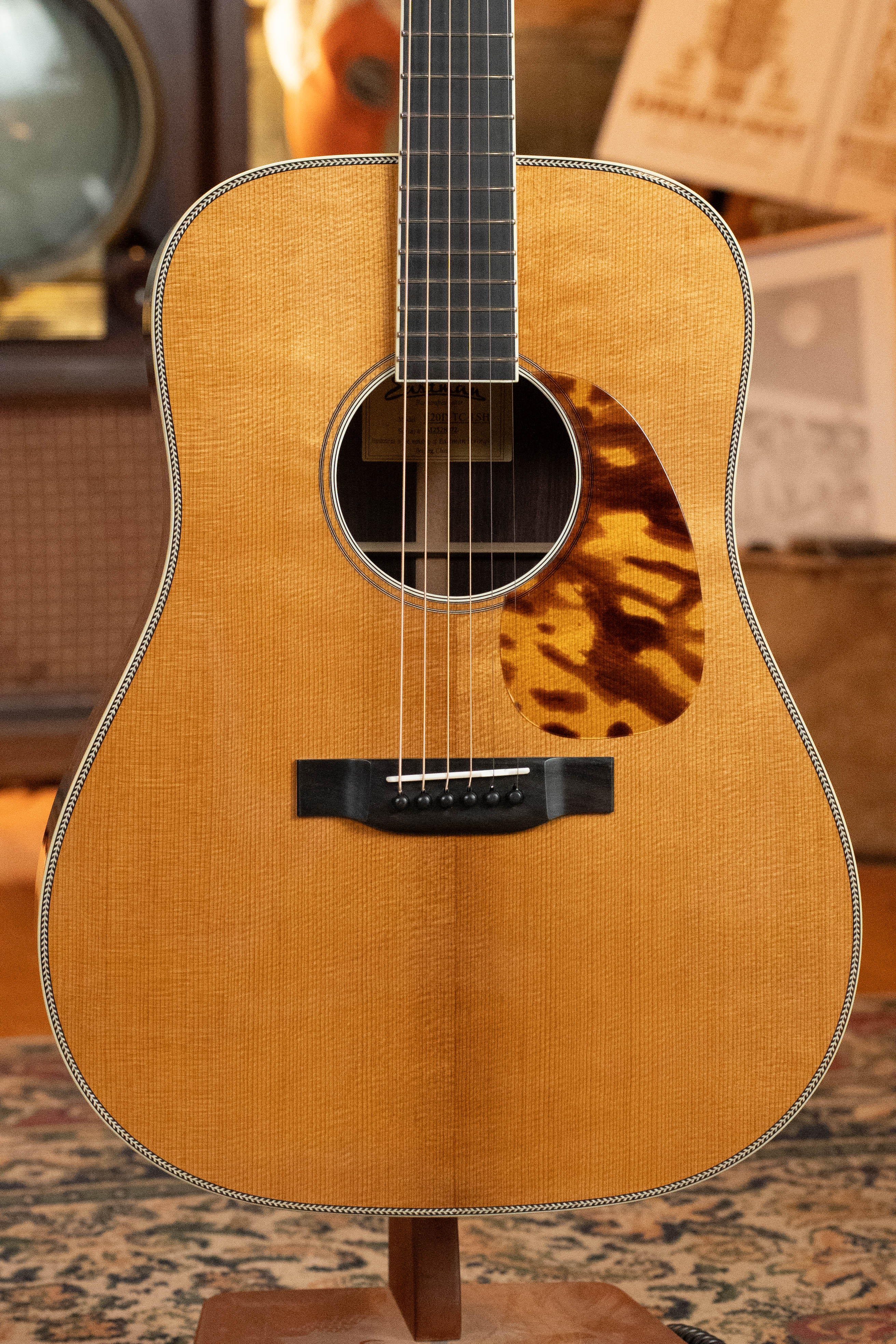 A close-up of an Eastman Guitars E20D-LSH-TC #8072 Dreadnought, featuring a thermo-cured Adirondack Spruce top and tortoiseshell pickguard, standing upright on a patterned rug with vintage decor in the background.