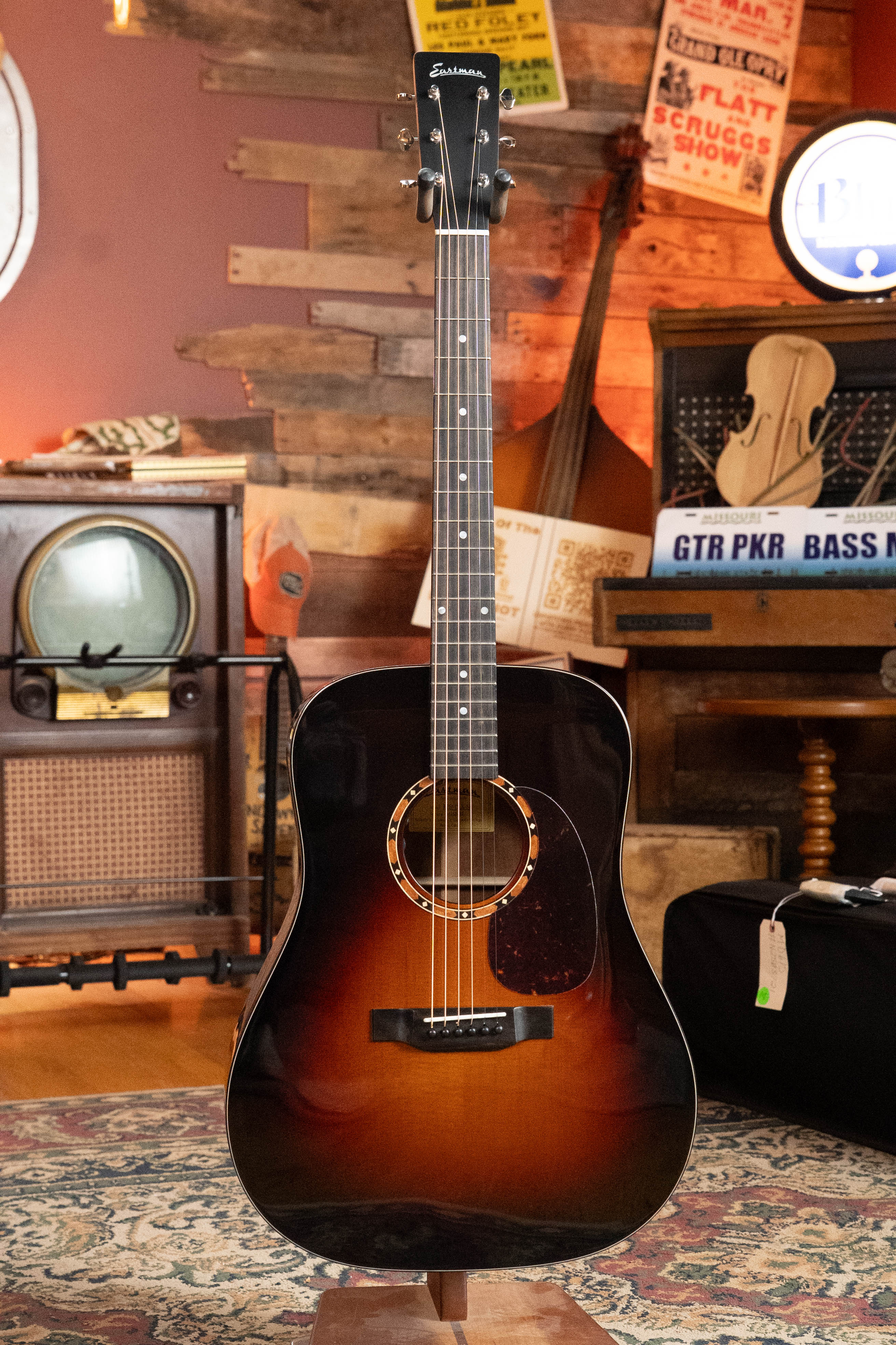 An Eastman E2D-DLX-SB Cedar/Sapele Sunburst Dreadnought Guitar with Fishman pickup by Eastman Guitars sits on a stand in a cozy, vintage room with wood walls, retro decor, a radio, and instruments in the background.
