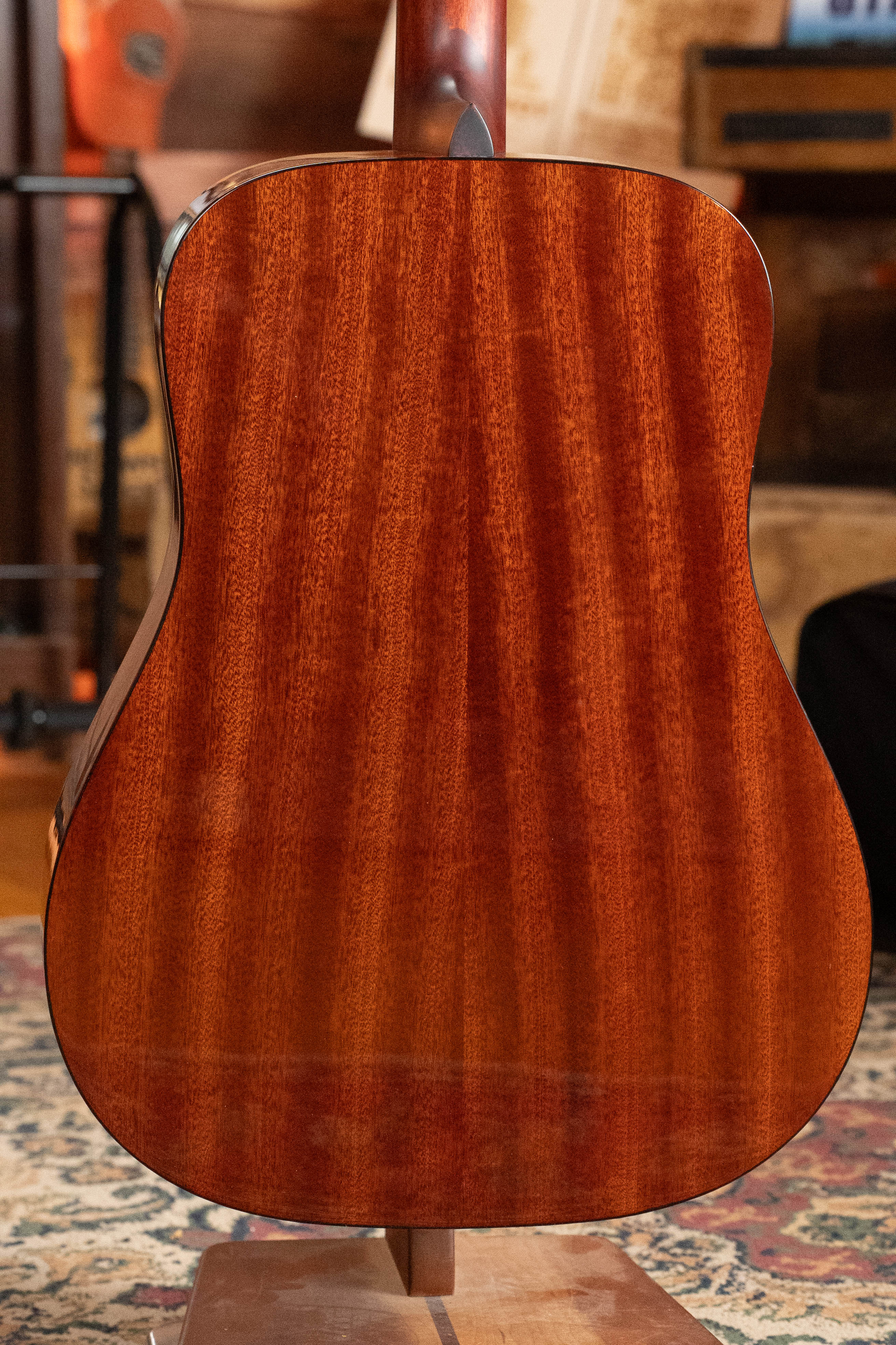 The image shows the back of an Eastman Guitars E2D-DLX-SB Cedar/Sapele Sunburst Dreadnought Guitar w/Fishman Pickup #1532, featuring polished reddish-brown wood grain, displayed on a wooden floor with a patterned rug and warm lighting.