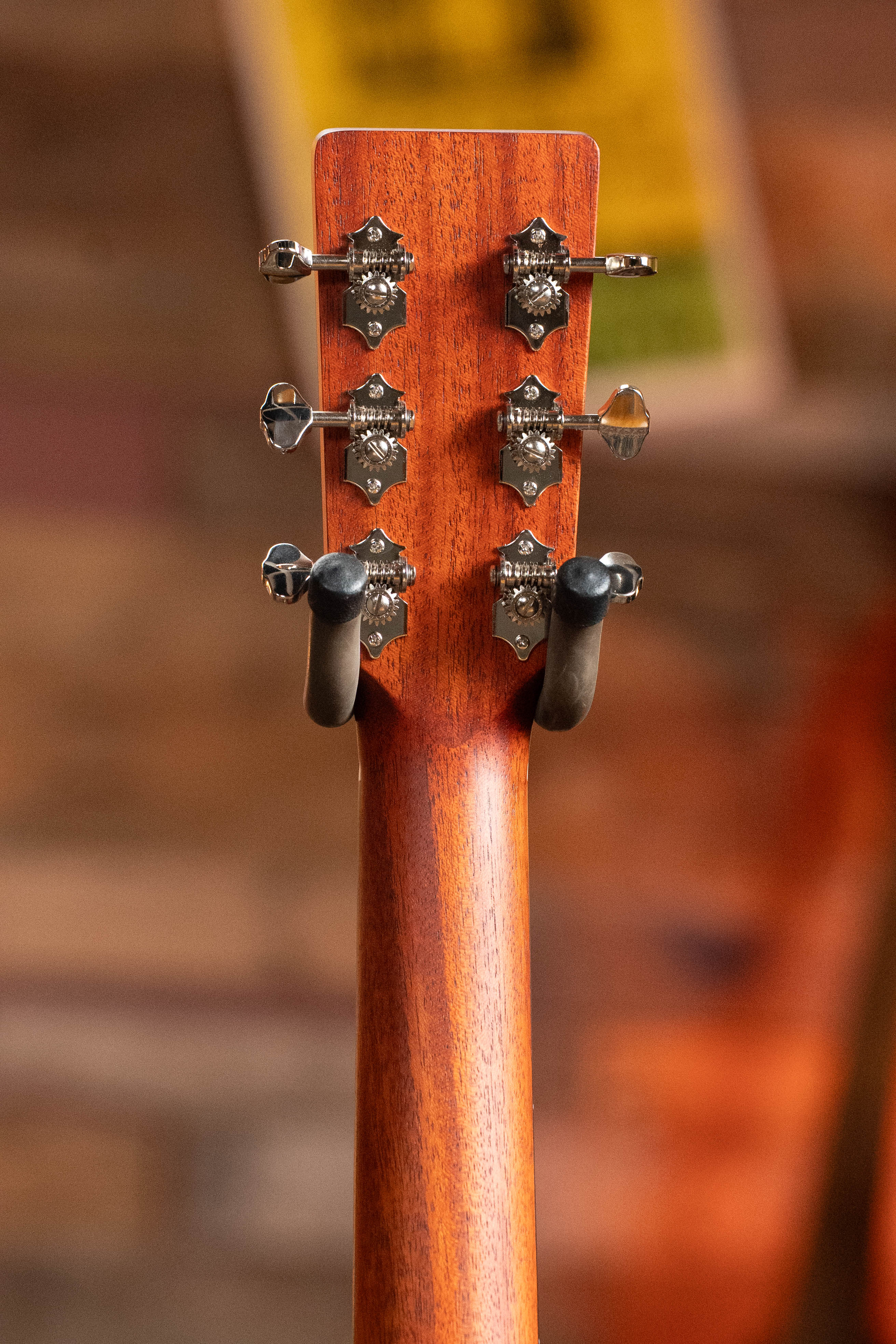 The image features the back of an Eastman Guitars E2OM Cedar/Sapele Orchestra Model Acoustic Guitar #9002 headstock with six tuning pegs and part of a wooden neck, set against a blurred, warm-toned background. Ideal for fingerstyle players.