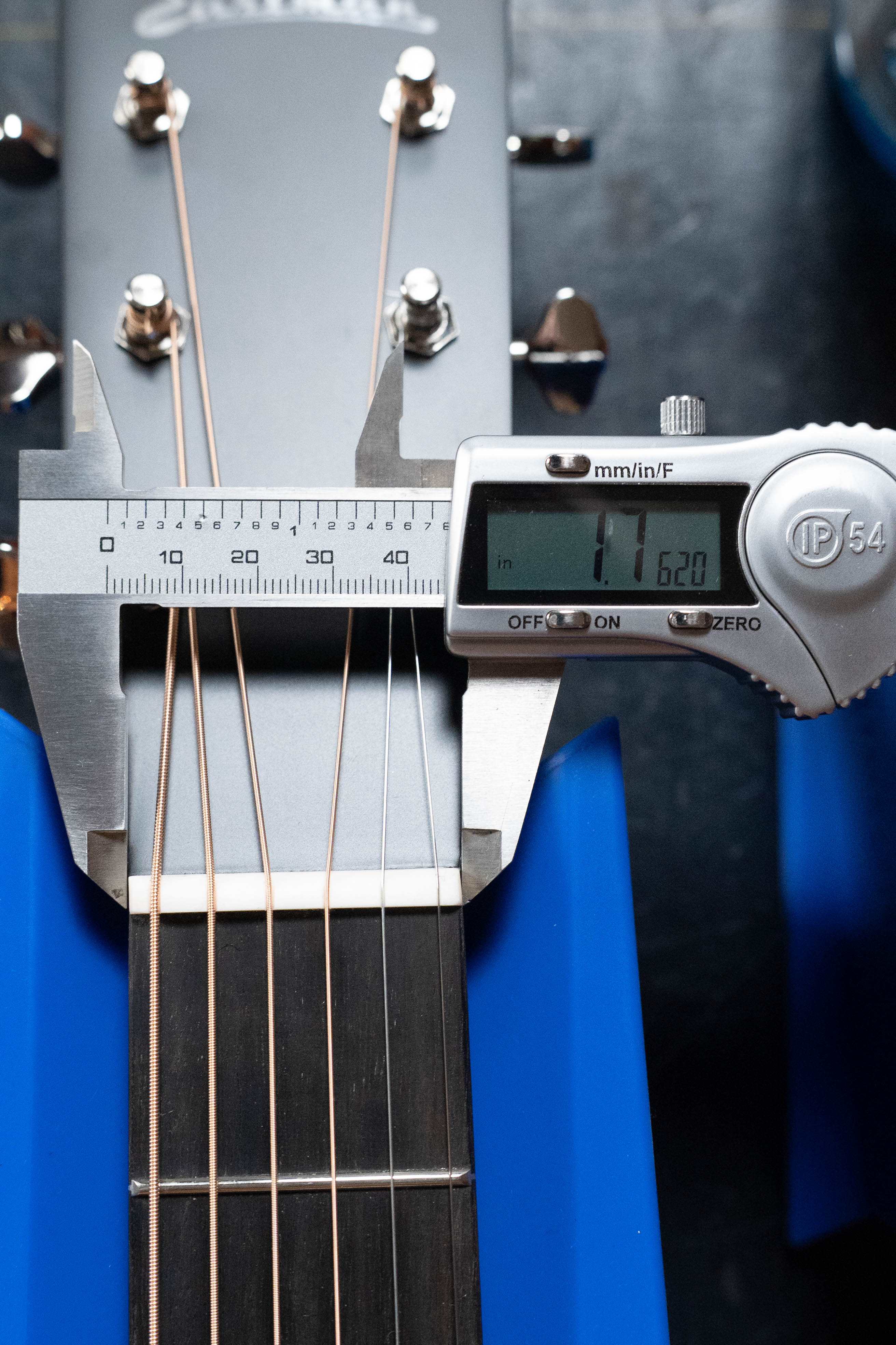 A digital caliper reads 1.620 inches as it measures the neck width above the nut of an Eastman Guitars E2OM Cedar/Sapele Orchestra Model Acoustic Guitar #9002, which features a solid cedar top and sits on a blue stand.