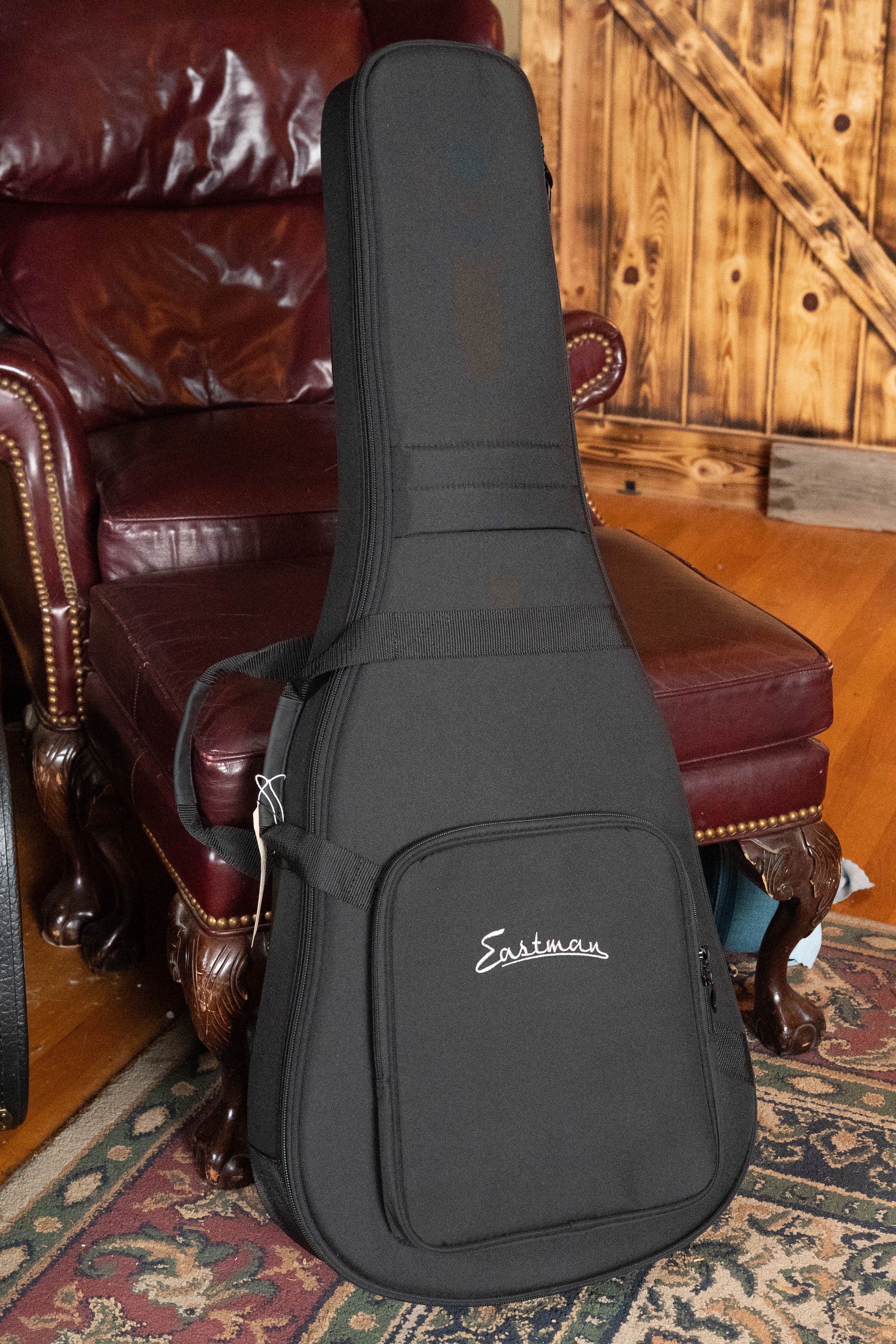 An Eastman Guitars E2OM Cedar/Sapele Orchestra Model Acoustic Guitar (#9002) in a black case rests upright against a burgundy leather armchair on a patterned rug, set in a room with wooden floors and a wooden door.
