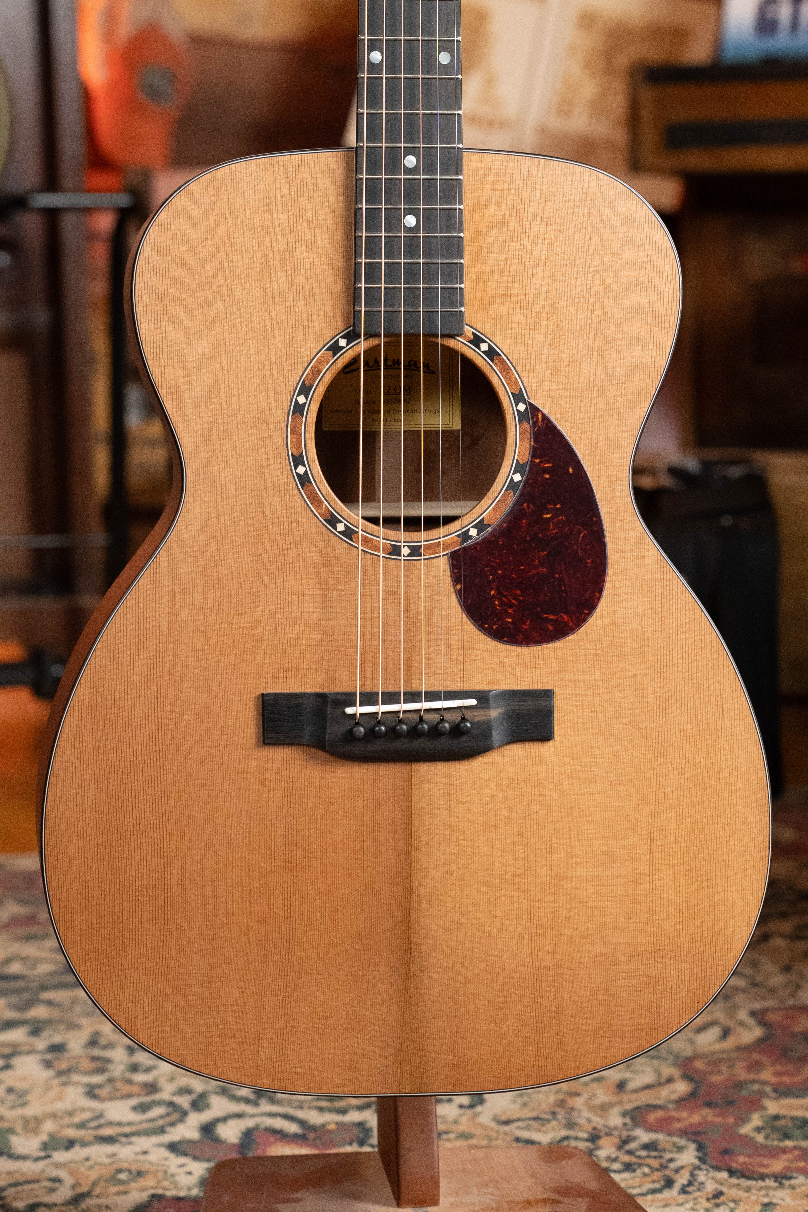 An Eastman Guitars E2OM Cedar/Sapele Orchestra Model Acoustic Guitar #9002 with a solid cedar top stands upright on a wooden stand in a cozy room with patterned rug and music gear—ideal for fingerstyle playing.