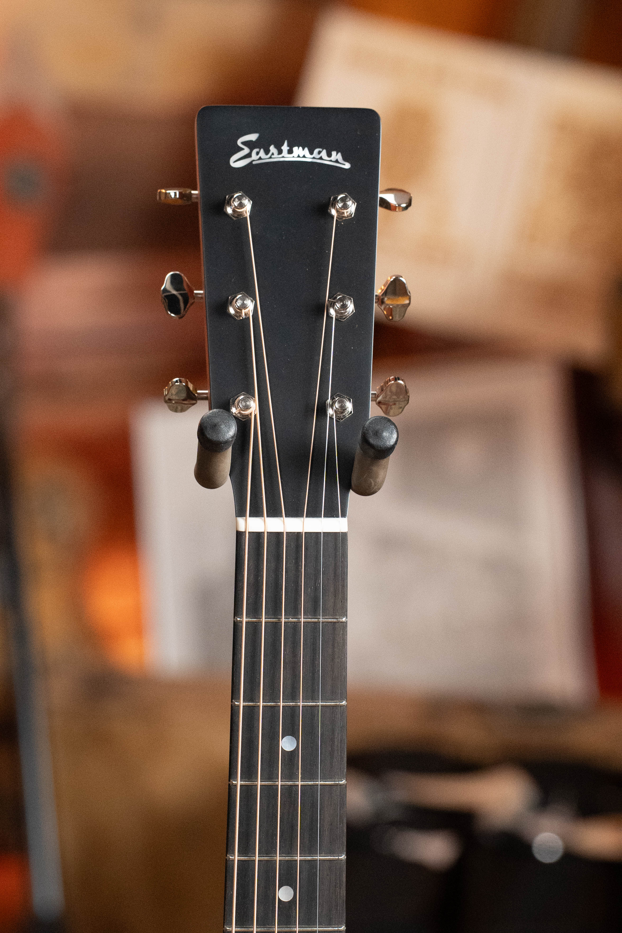 Close-up of the headstock on an Eastman Guitars E2OM Cedar/Sapele Orchestra Model Acoustic Guitar #9002, highlighting tuning pegs and strings, with a blurred background of wooden and paper objects.