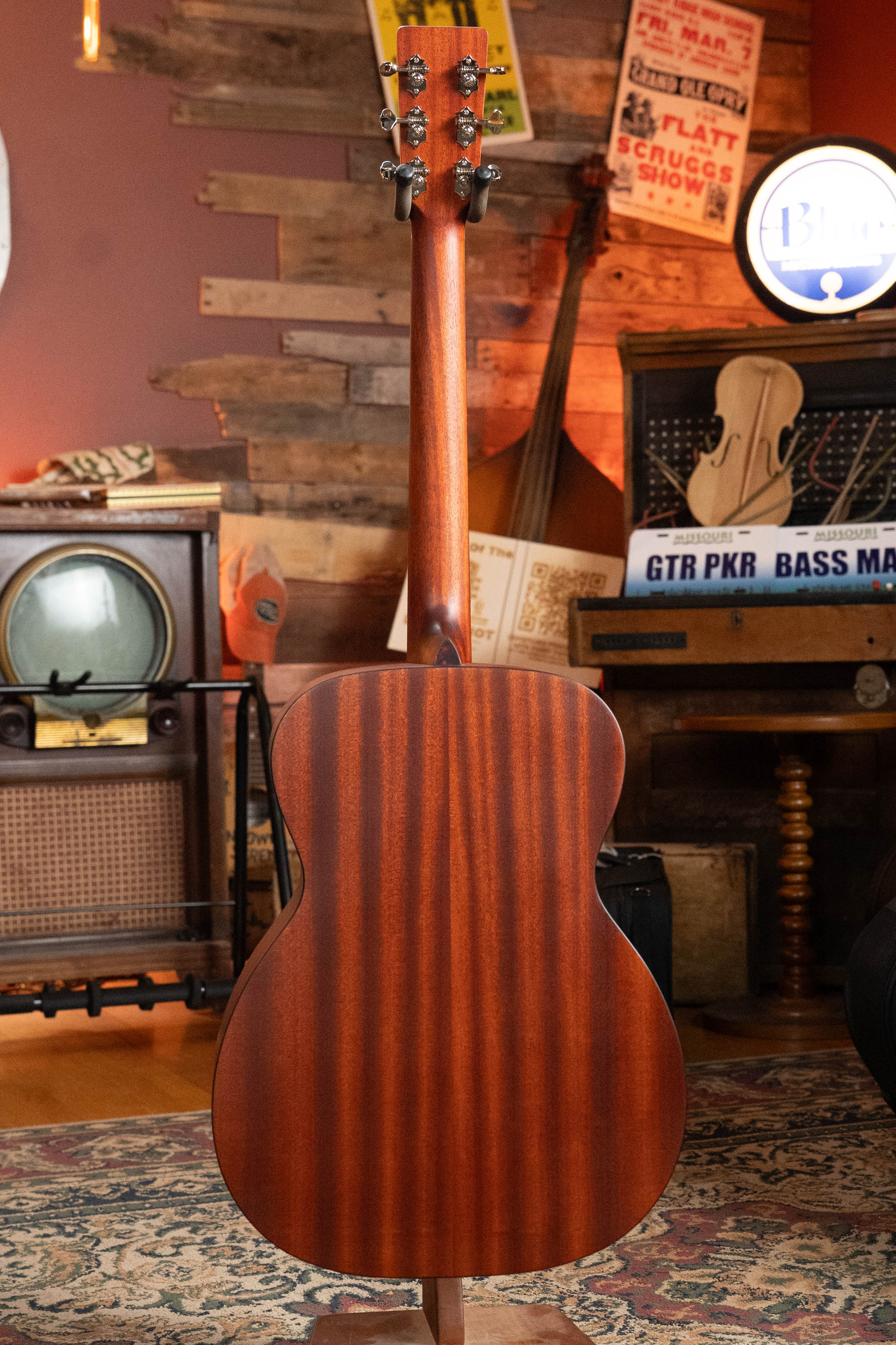 An Eastman Guitars E2OM Cedar/Sapele Orchestra Model Acoustic Guitar #9002 is displayed upright from the back in a cozy vintage room with retro décor, a rug, old TV, musical instruments, and nostalgic signs in the background.