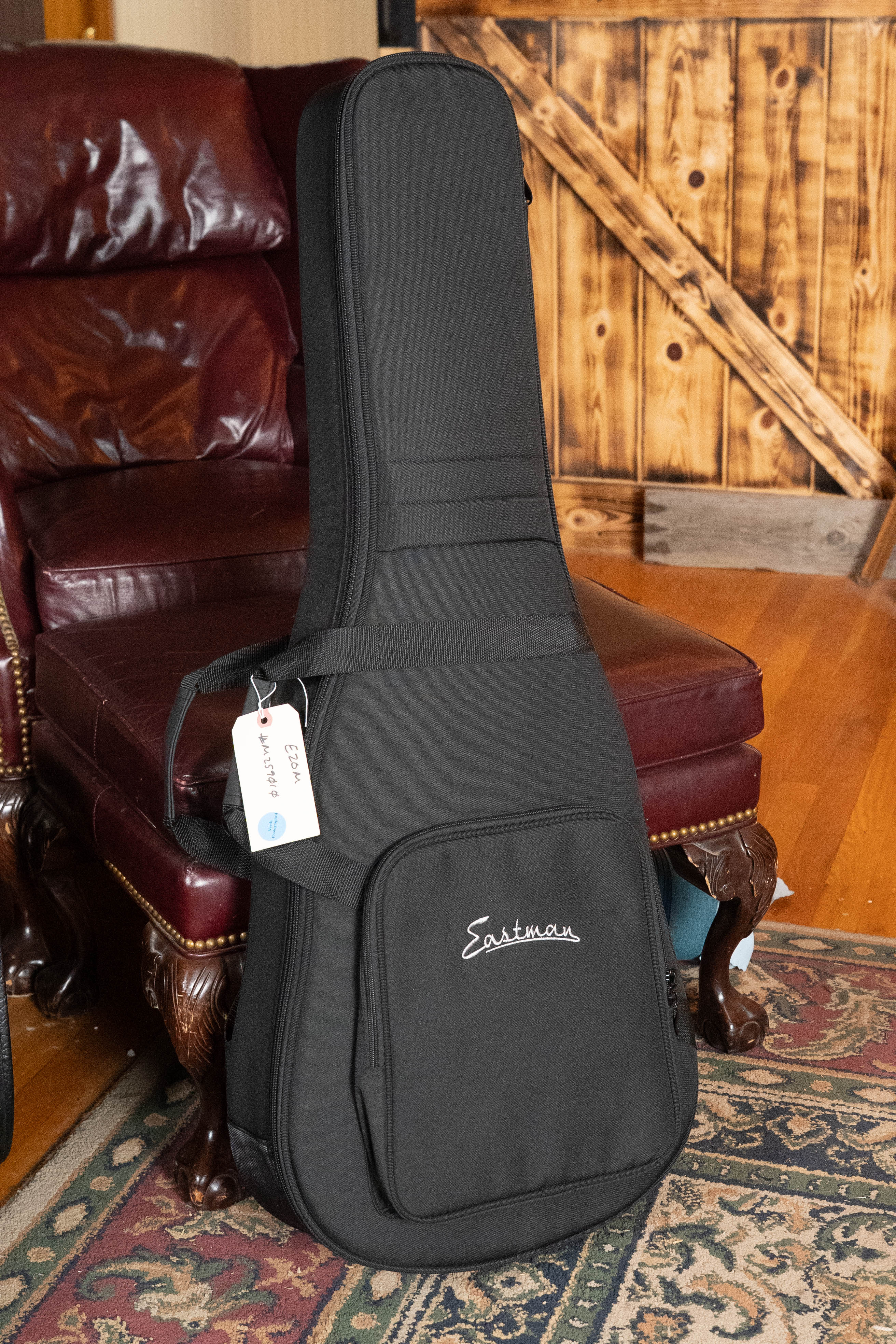 A black guitar case for the Eastman Guitars E2OM Cedar/Sapele Acoustic #9010, featuring a front pocket, carrying handle with white tag, sits on a patterned rug near a maroon leather chair and sliding barn door.