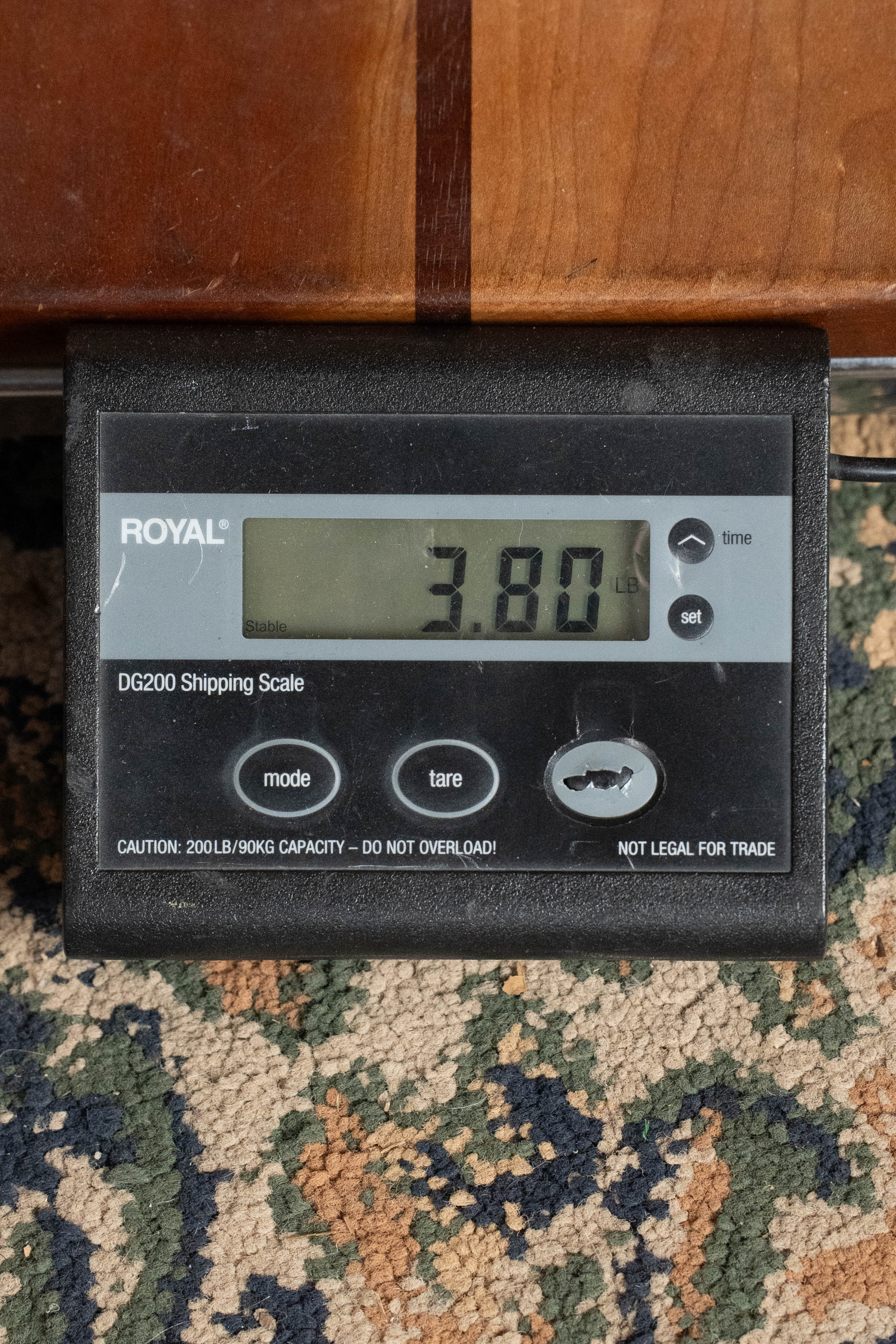 A digital shipping scale on patterned carpet shows 3.80 lbs—ideal for the Eastman Guitars E2OM Cedar/Sapele Orchestra Model Acoustic Guitar #9010. The scale features mode, tare, and power buttons with a cautioned capacity of 200 lbs. Perfect for fingerstyle players.