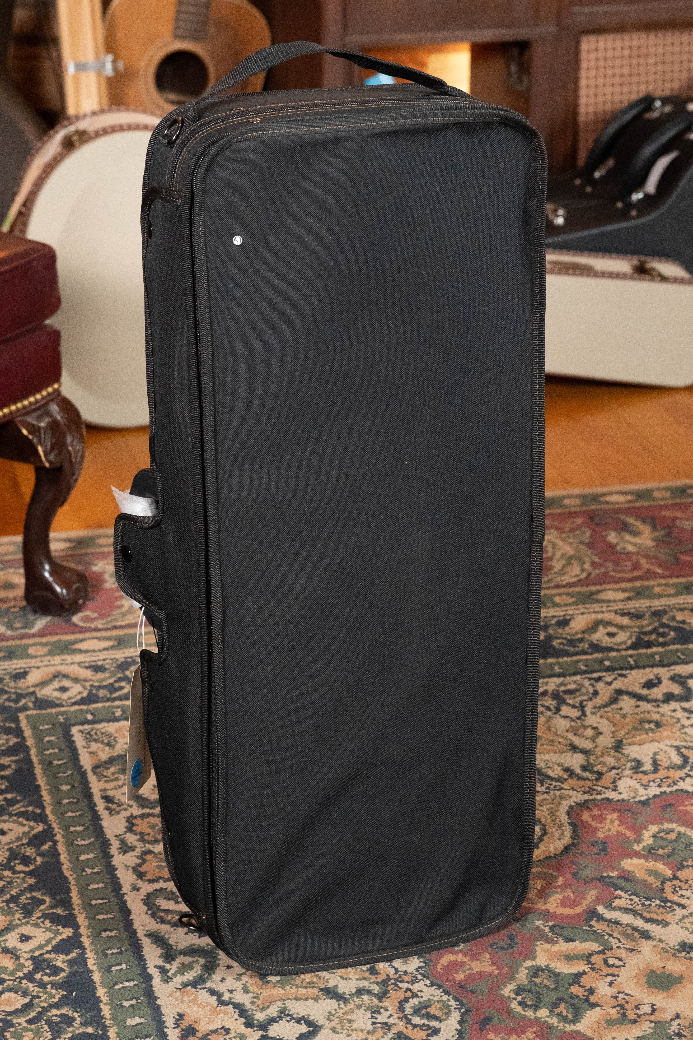 A tall, rectangular black carrying case stands upright on a rug indoors with a guitar, maroon chair, and an Eastman Guitars MD615 Spruce/Maple F-Style Mandolin w/K&K Pickup #3918 visible in the background.