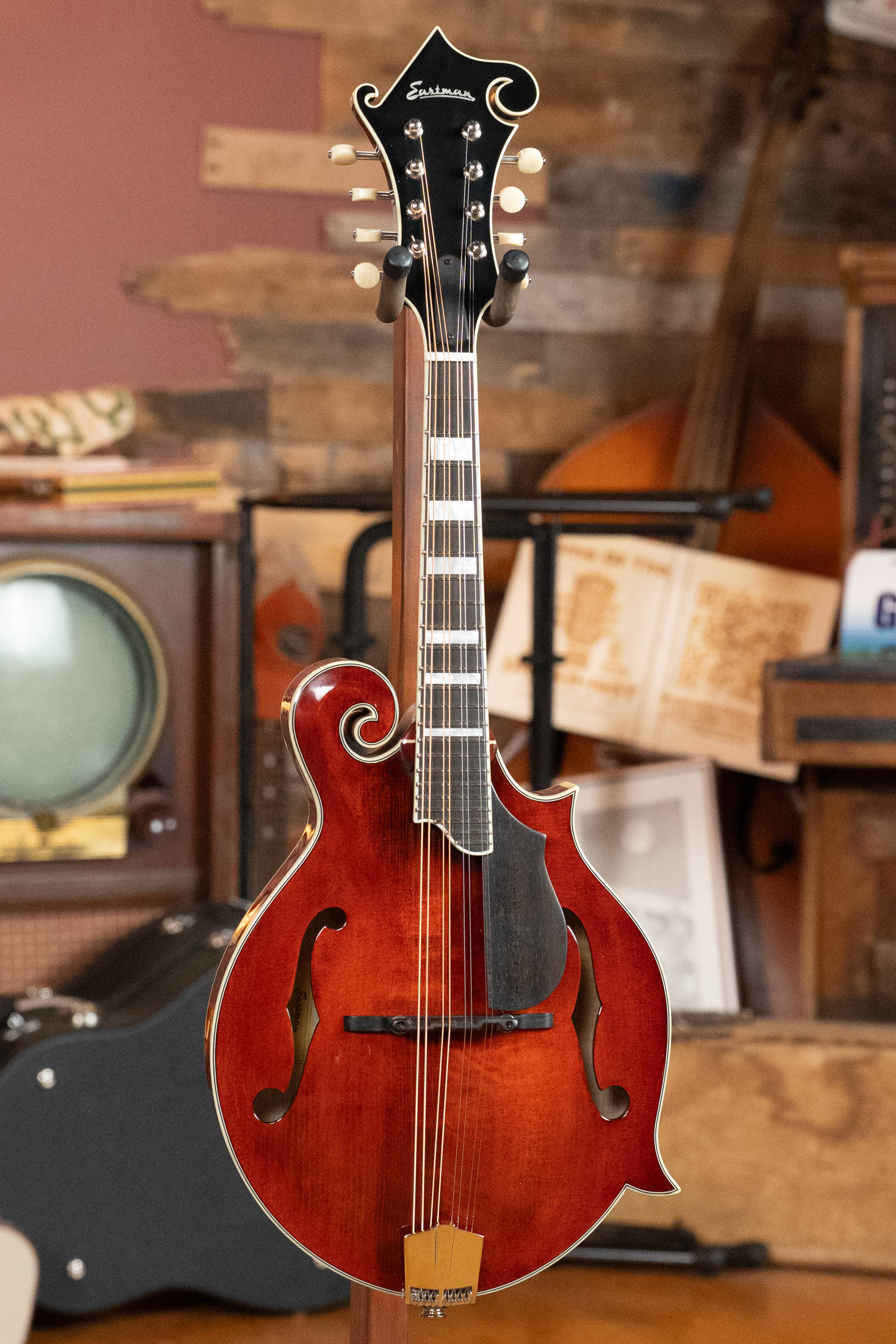 The Eastman Guitars MD615 Spruce/Maple F-Style Mandolin w/K&K Pickup #3918 is displayed upright indoors among vintage items, featuring a glossy red-brown finish, decorative headstock, and f-holes.