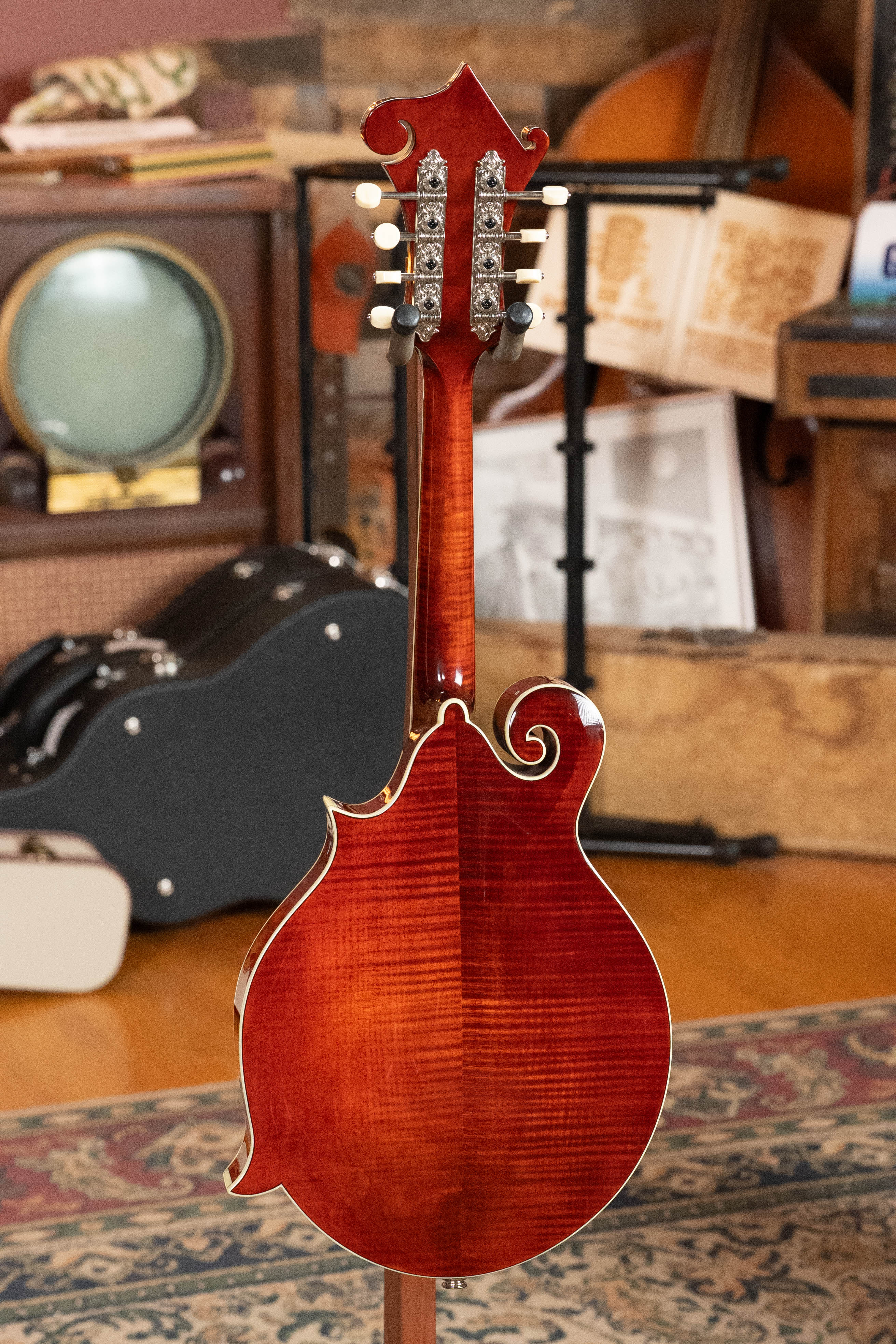The Eastman Guitars MD615 Spruce/Maple F-Style Mandolin w/K&K Pickup (#3918) is displayed from the back on a patterned rug, with musical instruments, a vintage TV, an open book, and a guitar case in the background.