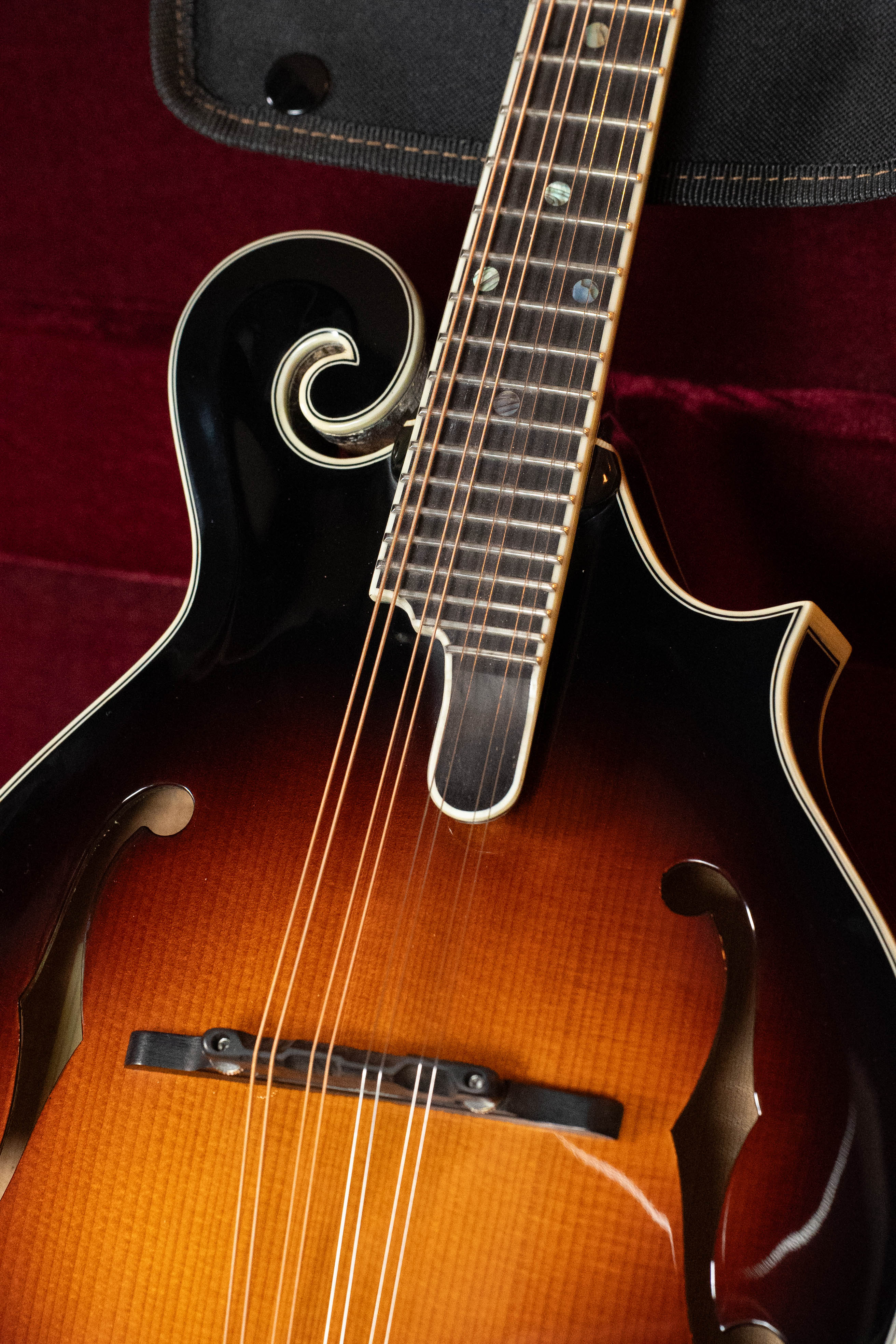Close-up of an Eastman Guitars MD815-SB Adirondack/Flamed Maple Sunburst F-Style Mandolin #3355, showing its elegant curves, decorative scroll, and f-holes against a deep red background.