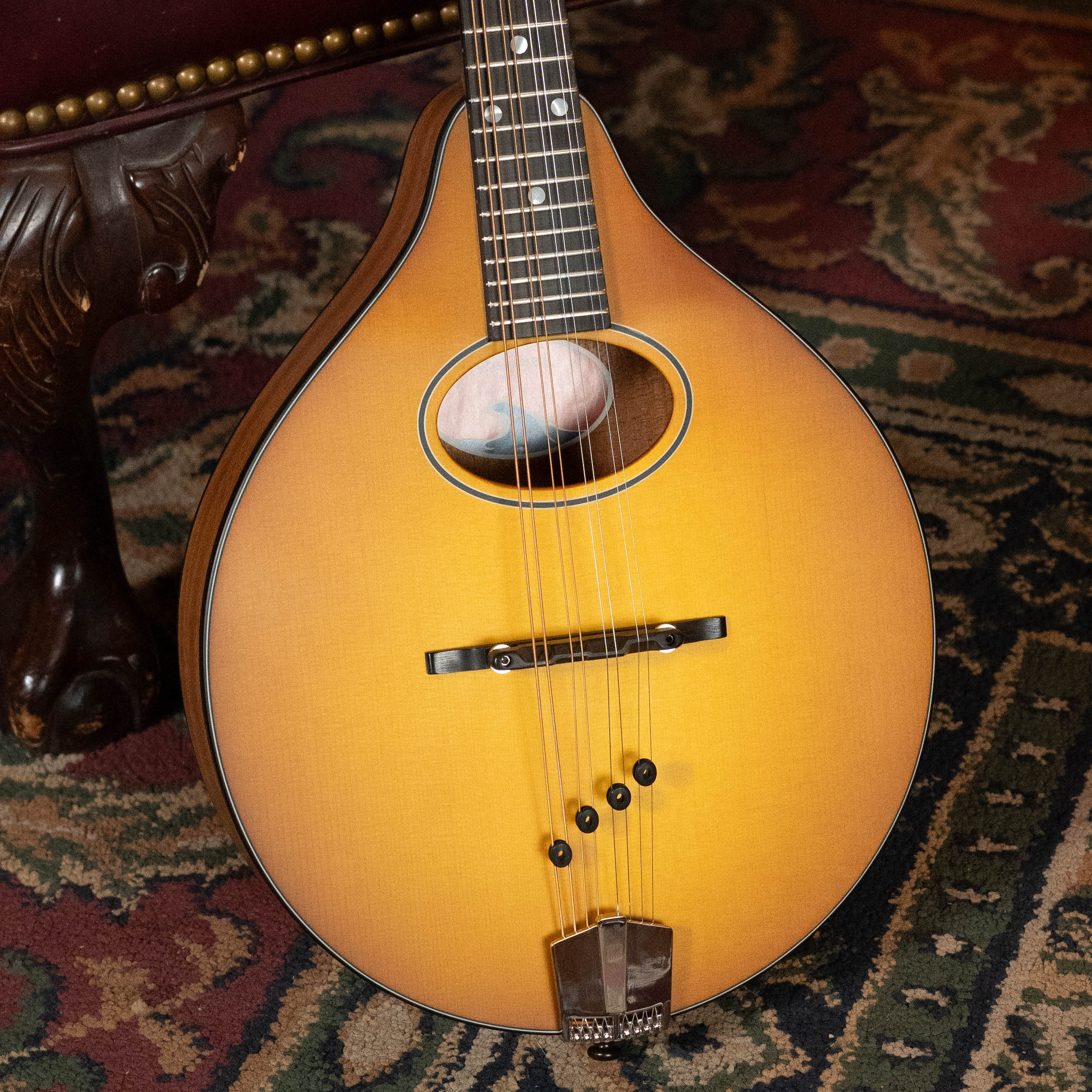 A close-up of the Eastman Guitars PCH-M104-GB Solid Spruce/Laminated Mahogany A-Style Mandolin #0053 in goldburst, resting on a patterned carpet by a wooden chair leg—an excellent choice for beginner mandolin players.