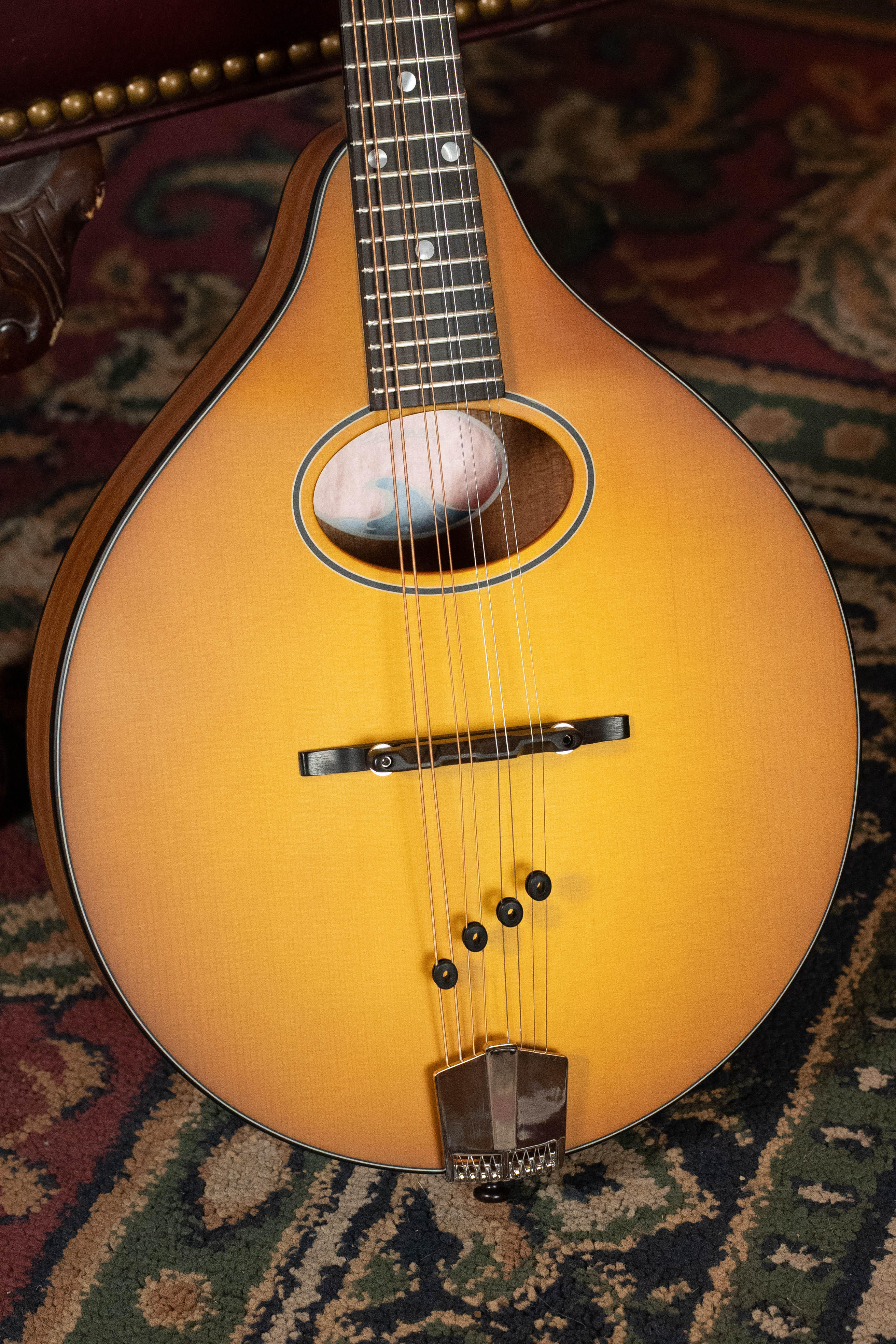 A close-up of the Eastman Guitars PCH-M104-GB Solid Spruce/Laminated Mahogany Goldburst Flattop A-Style Mandolin #0053 on a patterned rug, highlighting its strings, sound hole, and neck—perfect for beginner players.