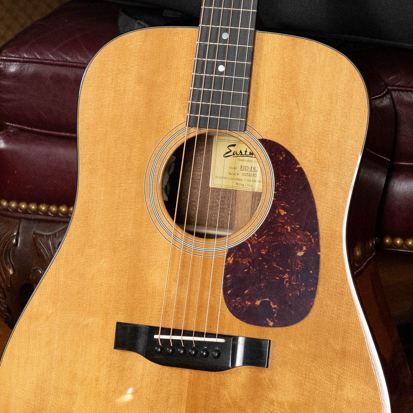 A close-up of the Eastman Guitars E1D-DLX Sitka/Sapele Dreadnought w/Fishman Pickup #2371 on a maroon leather chair, highlighting its strings, soundhole, pickguard, and part of the inside label.